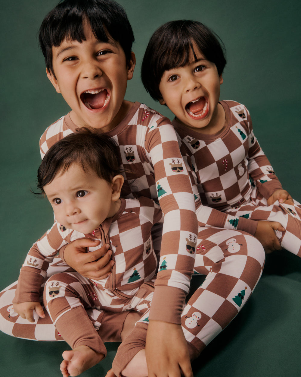 Three siblings wearing matching Holiday Checks PJs on a green backdrop