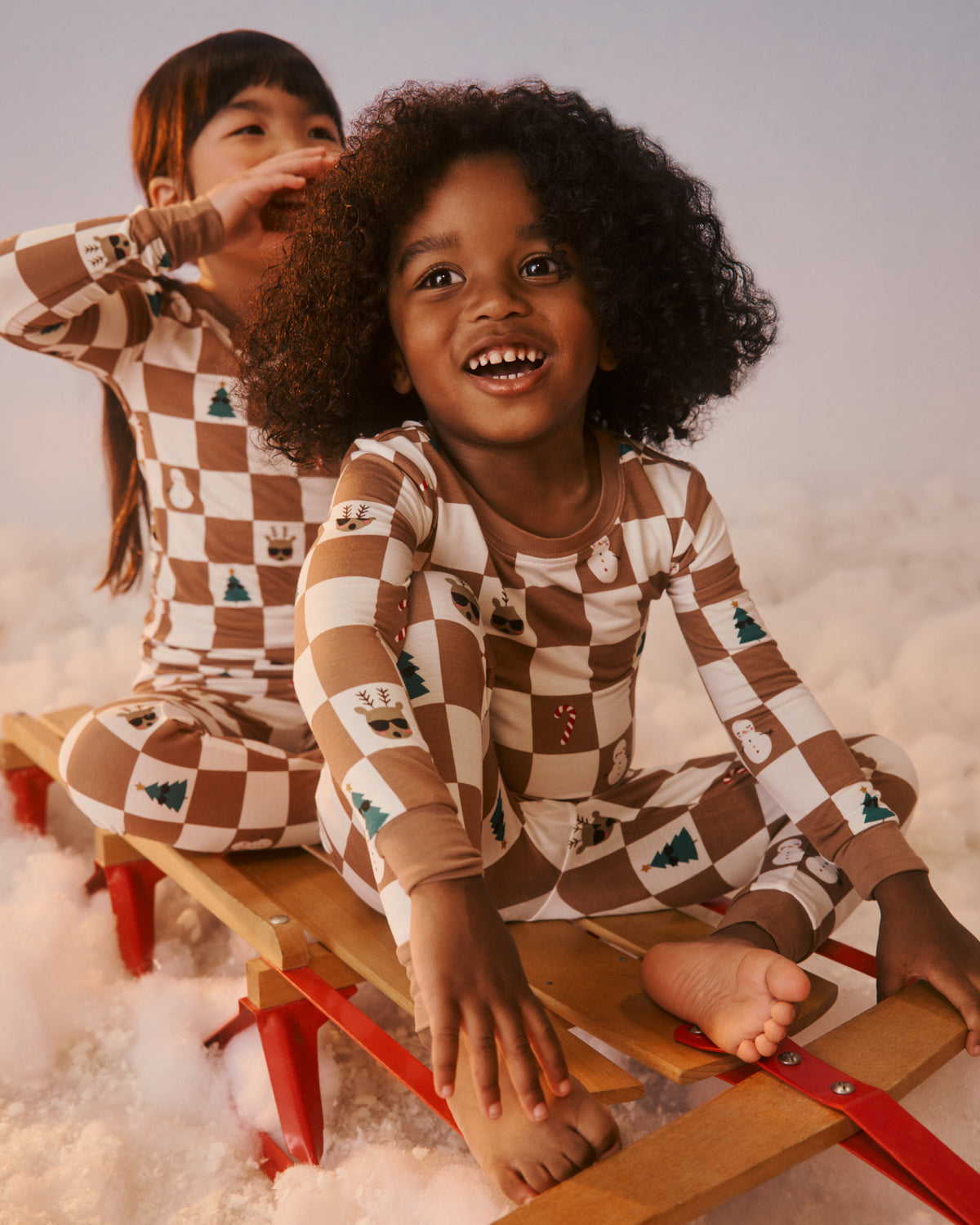 Two children wearing matching brown and cream checkered pajamas with small holiday graphics, sitting on a wooden sled in the snow.