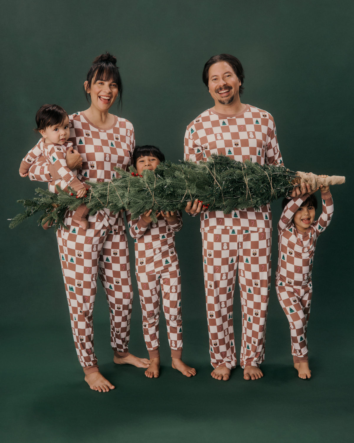 A family of five wearing matching checkered pajamas stands barefoot against a dark green studio background, holding a large Christmas tree. The pajamas have a brown and beige checkerboard pattern with small holiday motifs.
