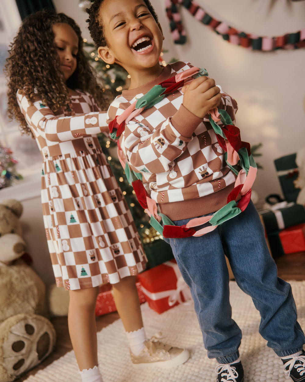 Two children wearing matching brown and white checkered holiday-themed long-sleeve shirts and dresses in a festive Christmas room setting with a tree and wrapped gifts.