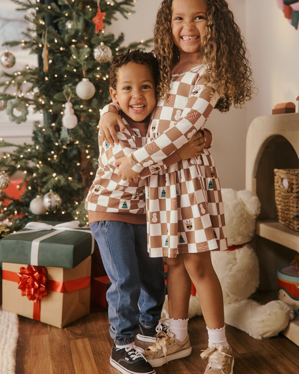 Two children wearing matching brown and cream checkered holiday styles standing in front of a decorated Christmas tree, with wrapped gifts in a cozy indoor setting.