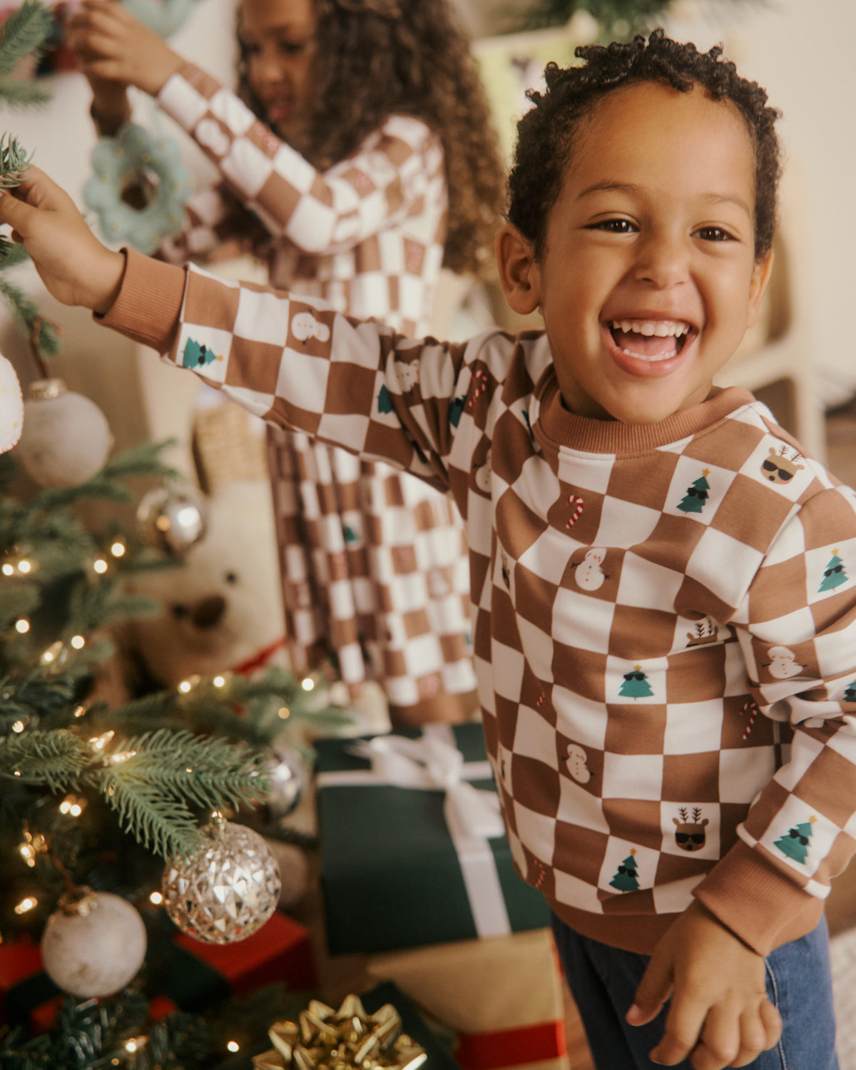 A young boy smiling and decorating a Christmas tree, wearing a brown and white checkered holiday pajama shirt with small tree and snowman motifs. A girl in a matching dress is visible in the background.