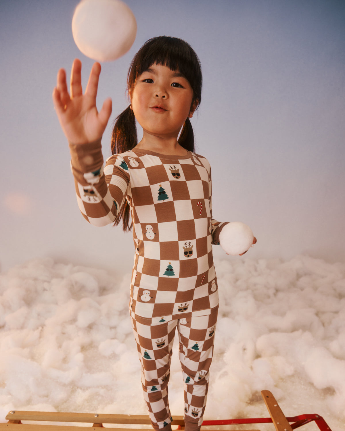 A young child wearing a brown and cream checkered one-piece pajama with small holiday motifs, standing on a wooden sled in a snowy studio setup.