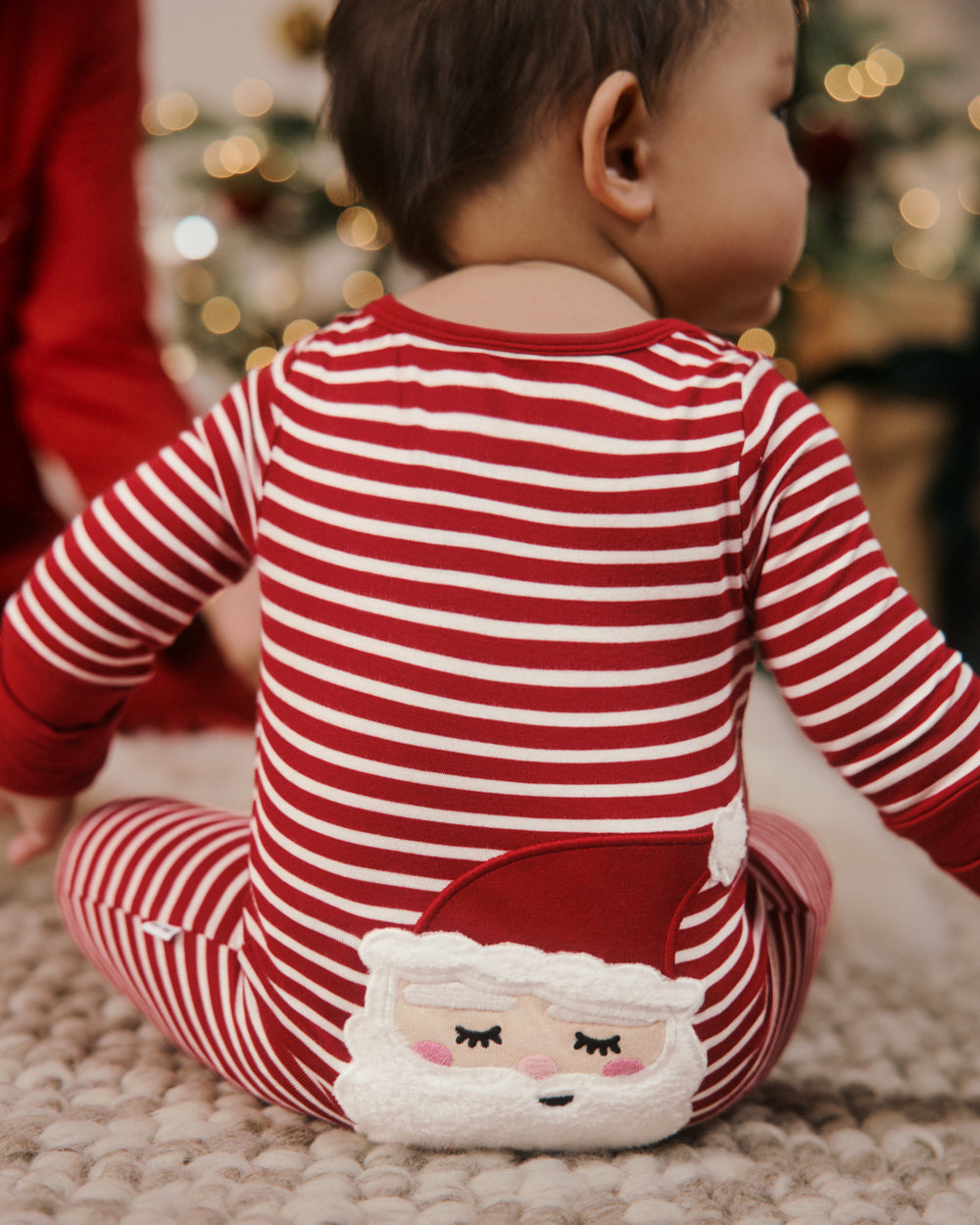 A baby sitting on a textured cream rug, viewed from behind, wearing a red and white striped long-sleeve pajama with a plush Santa face on the back.