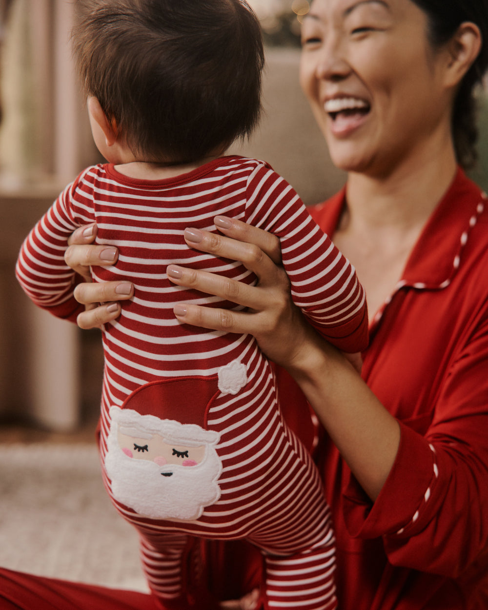 A baby wearing a red and white striped Christmas-themed footless one-piece pajama with a Santa face graphic on the back, held by a smiling woman in matching red pajamas.