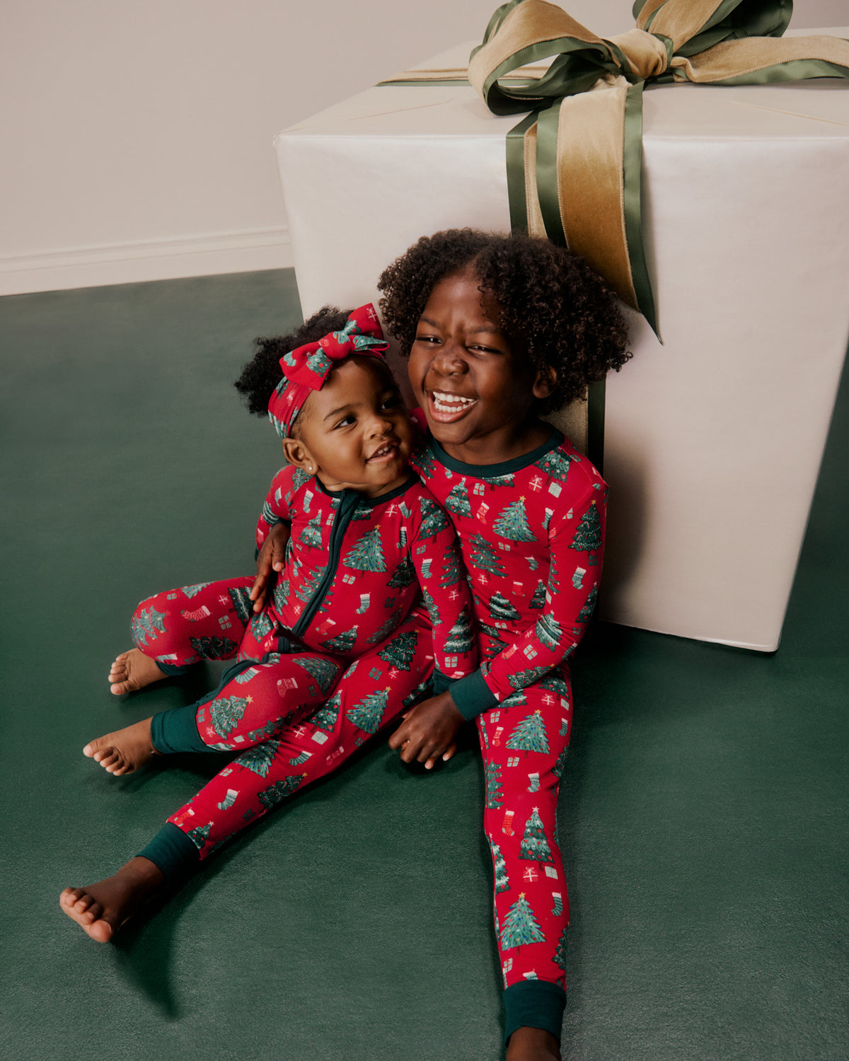 Two young kids wearing matching red Christmas-themed pajamas with a green tree pattern, sitting on a dark green floor next to a large wrapped gift box with a gold ribbon.