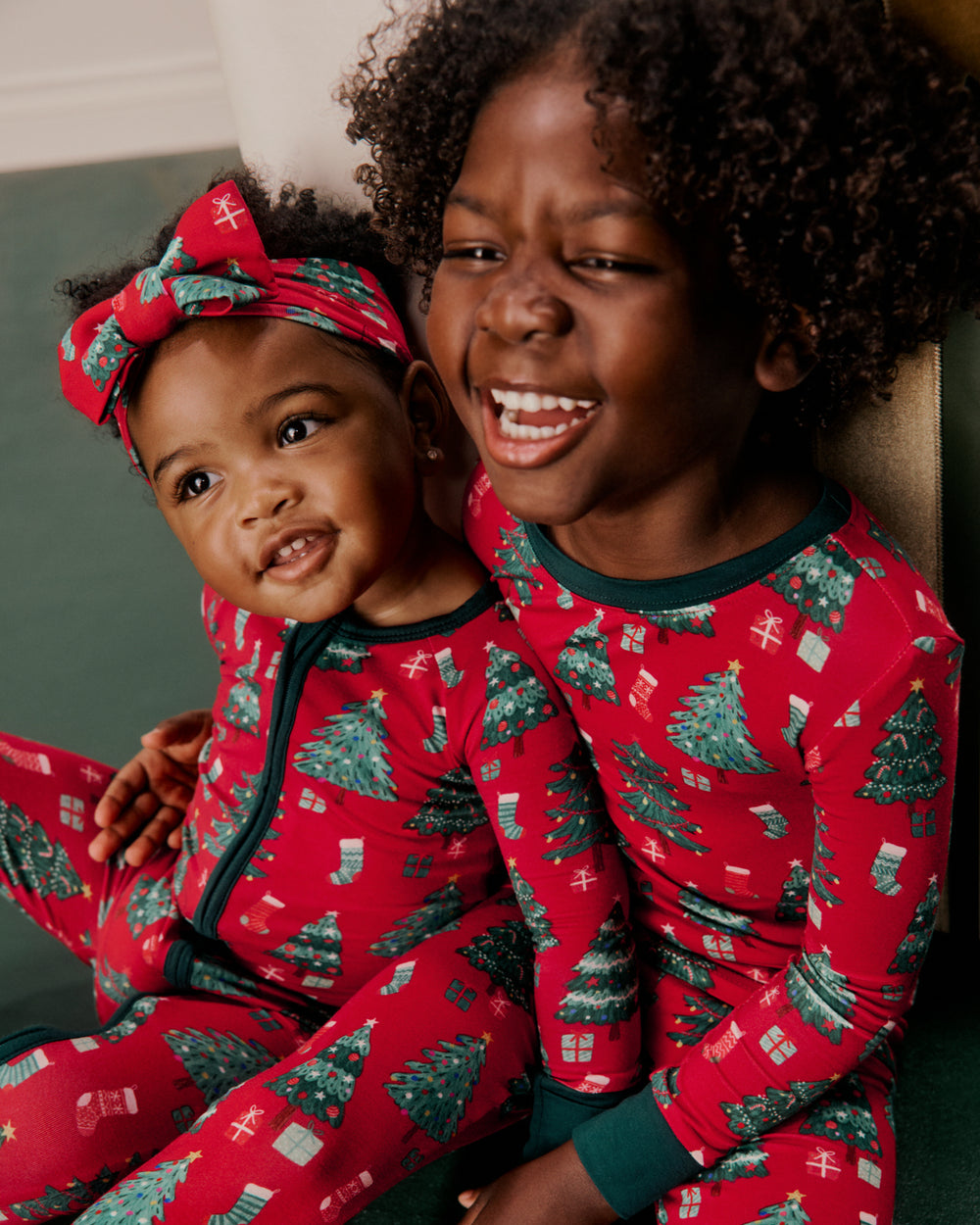 Two children wearing matching red Christmas-themed pajamas with a green tree print, sitting together indoors.