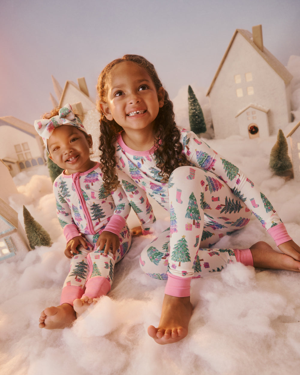 Two young girls sitting on fluffy white ground, wearing matching holiday-themed pajamas with a pink and white print of Christmas trees, houses, and stockings. 