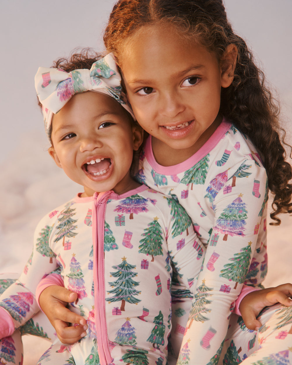 Two young girls wearing matching holiday-themed pajamas with a Christmas tree and stocking print, posed together against a soft light background.