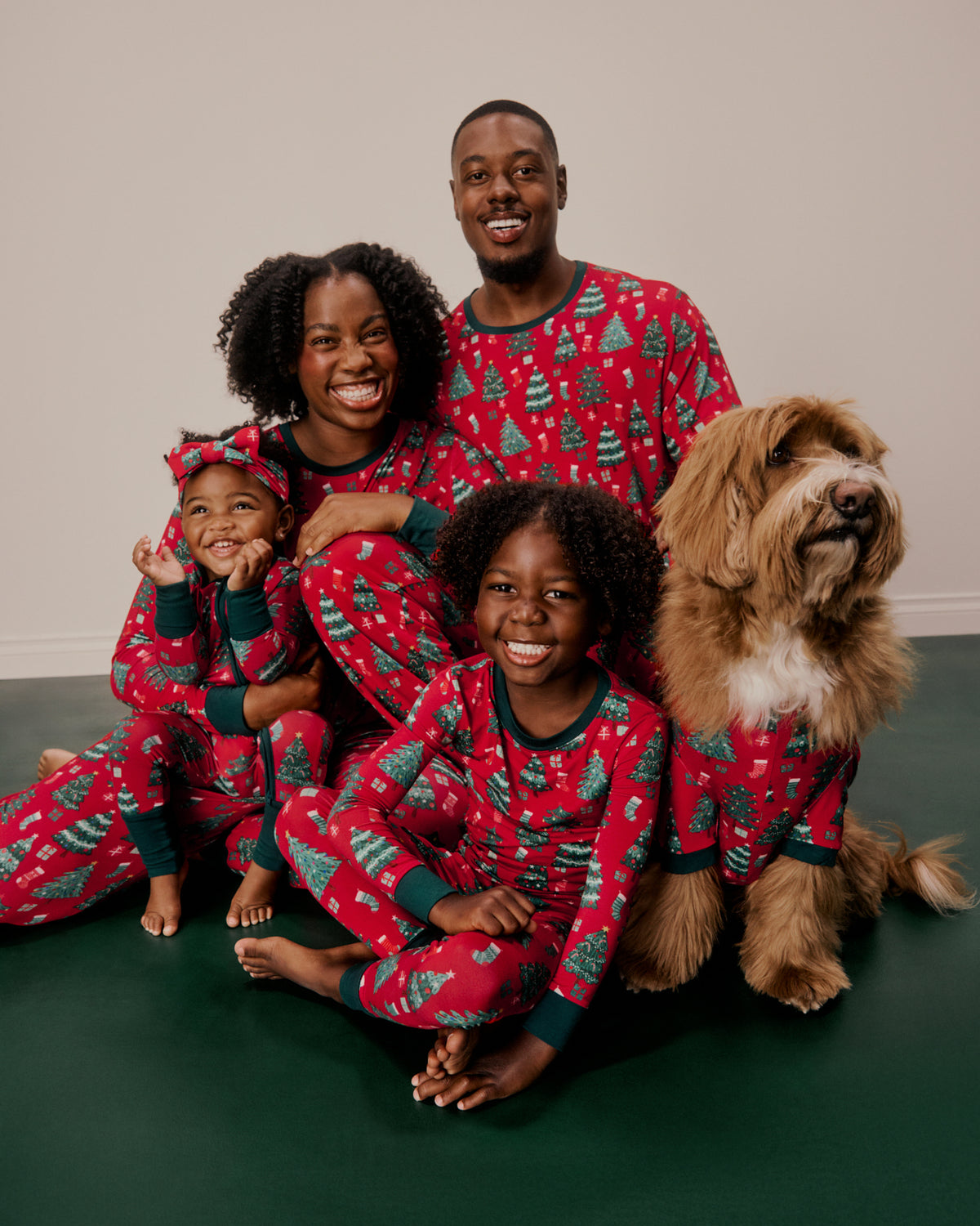 A family of four and their dog wearing matching red Christmas pajamas with a green tree pattern, posed together on a dark green floor against a light beige wall.