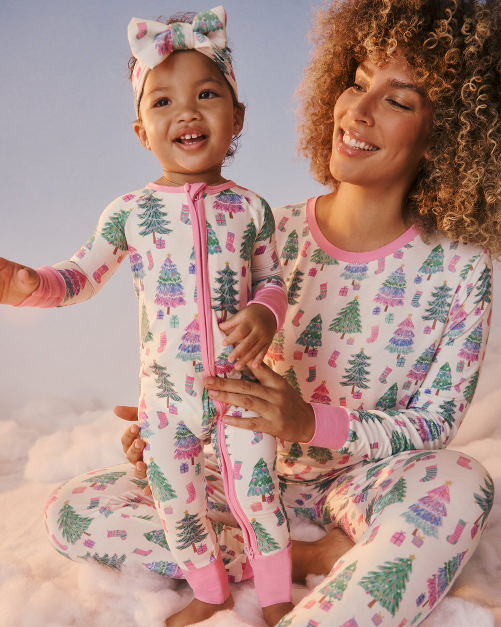 A smiling woman and child wearing matching pajamas with a Christmas tree and gift print, sitting together on fluffy white clouds against a soft blue sky. The pajamas are off-white with pink trim and a multicolor holiday pattern.