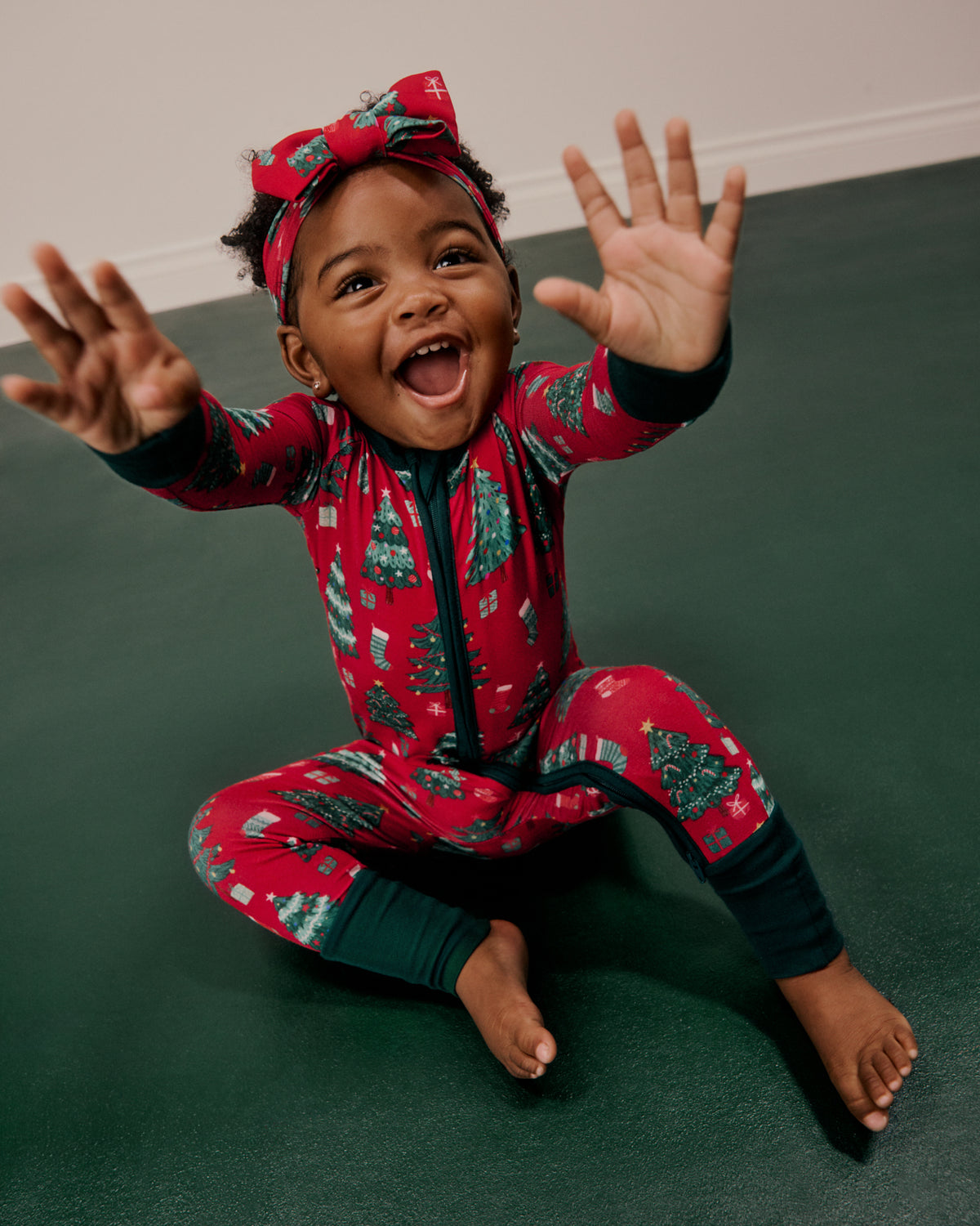 A smiling toddler sitting on a dark green floor wearing a festive red Christmas-themed one-piece pajama with a matching bow headband.
