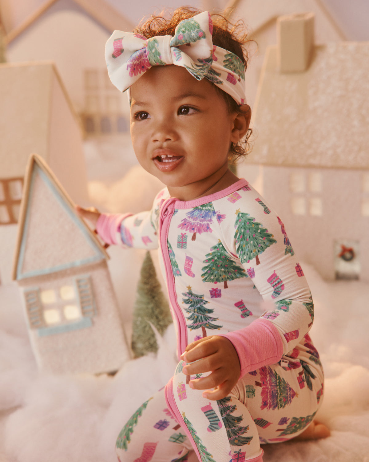 A toddler wearing a festive Christmas-themed one-piece pajama and matching bow headband, sitting on a soft white surface with toy houses in the background.