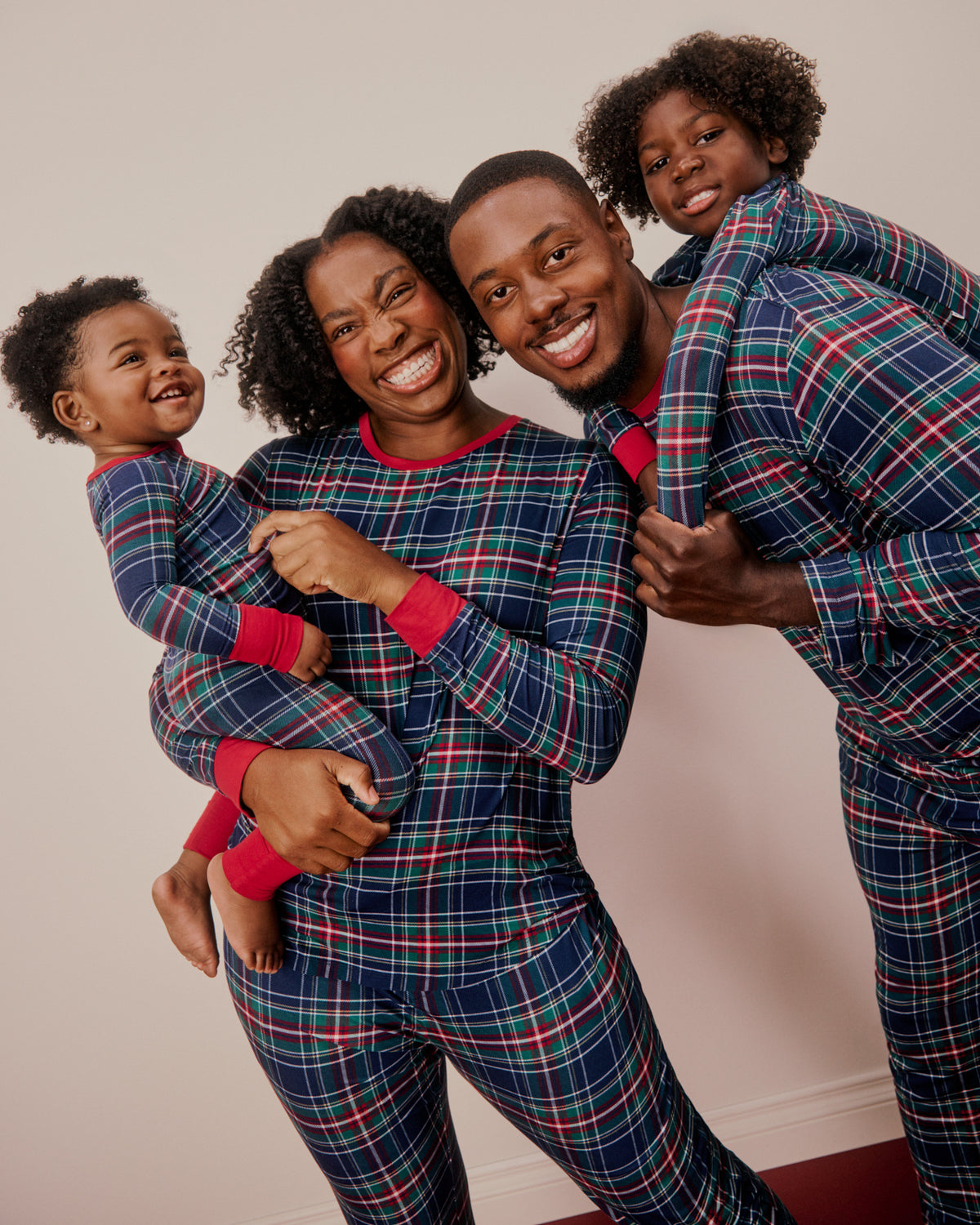 A family of four wearing matching plaid pajamas poses together against a plain beige wall. The pajama set features a navy blue base with red and green plaid lines and bright red cuffs and collar trim.