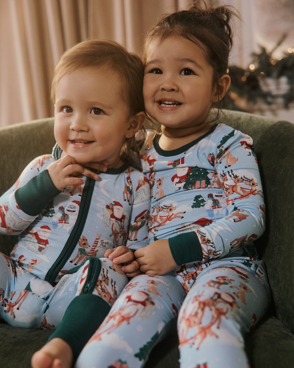 Two young children sitting together on a green couch, wearing matching light blue Christmas-themed pajamas with a festive print and dark green cuffs and zipper trim.