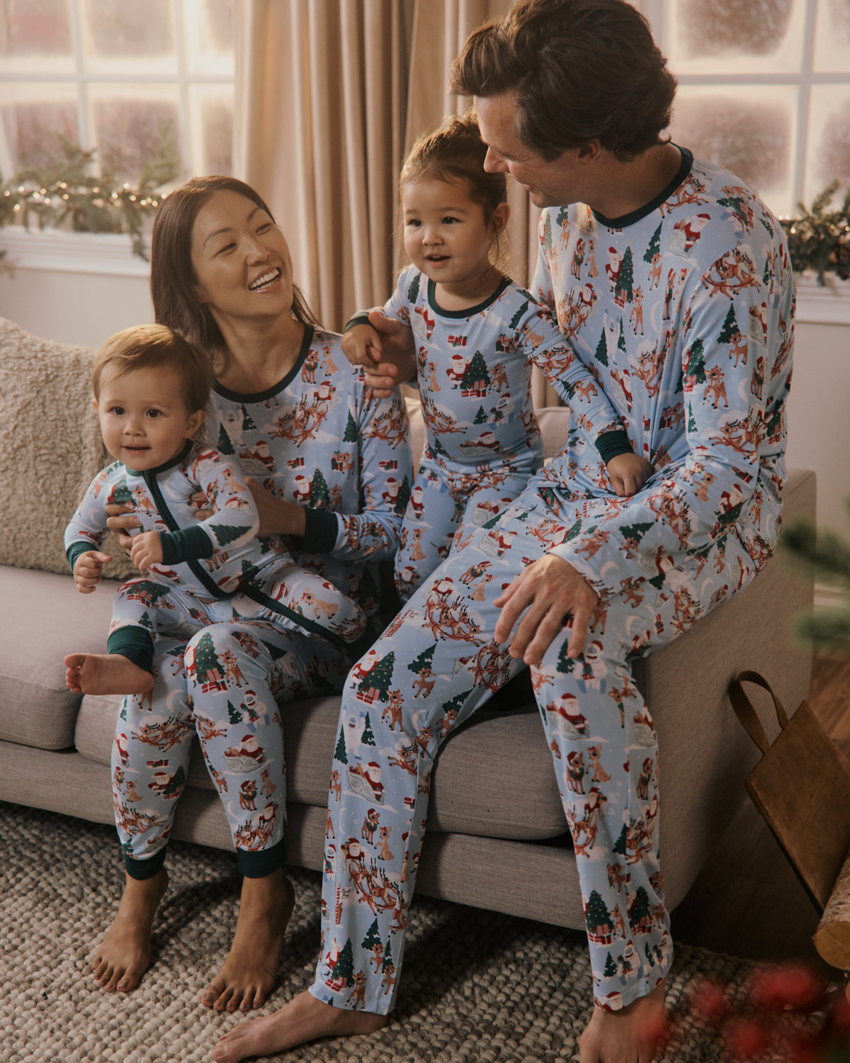 A family of four wearing matching light blue Christmas-themed pajamas with a festive print, sitting together on a couch in a cozy living room.