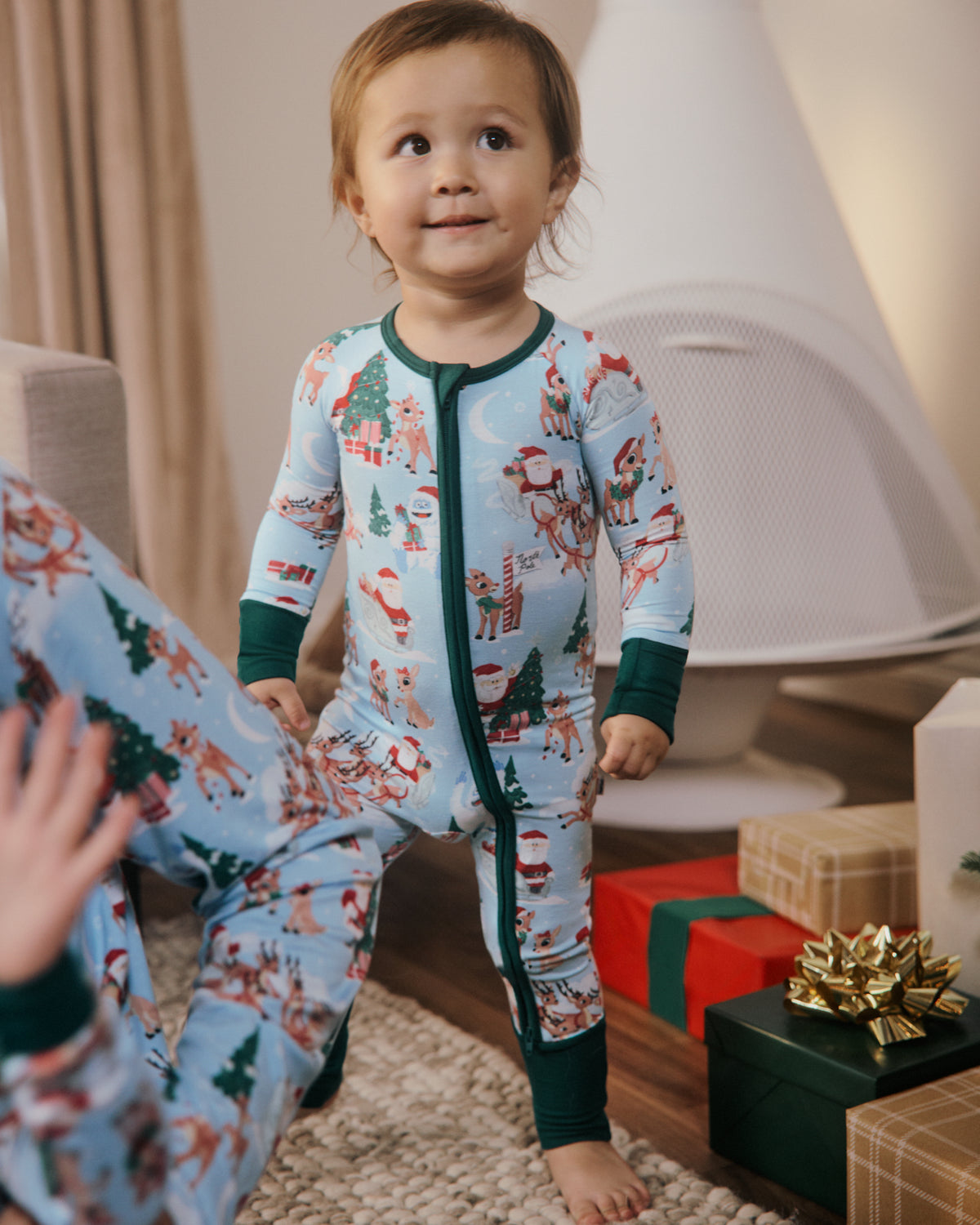 A toddler stands barefoot indoors wearing a light blue Christmas-themed one-piece pajama with green trim and a festive print of Santa Claus, trees, and gifts. A parent is seated at the left edge, and wrapped holiday presents are visible on the floor in a cozy living room setting.