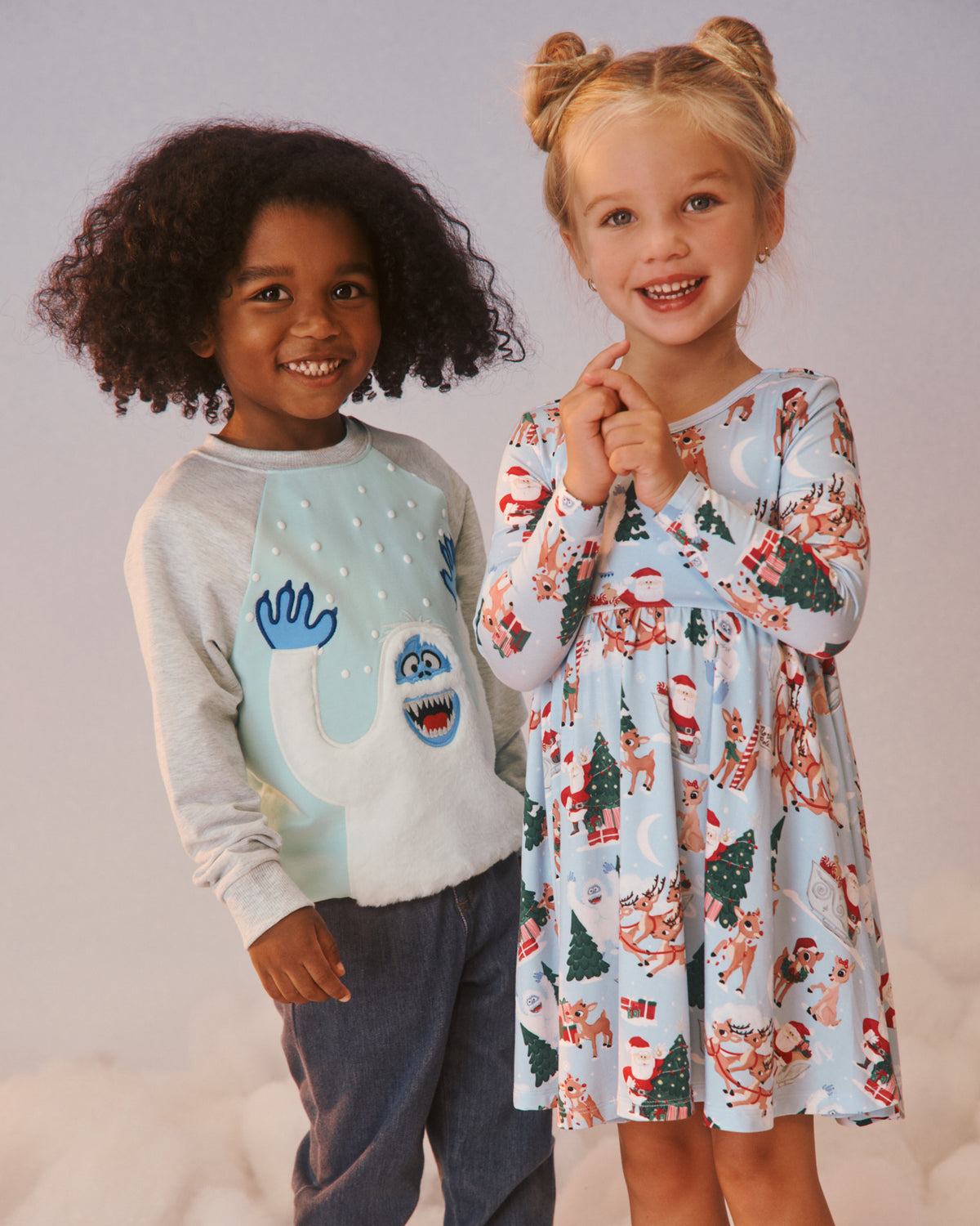 Two children modeling holiday-themed clothing against a soft light gray studio background. The child on the left wears a light gray and pale blue raglan sweatshirt with a blue monster graphic, paired with dark pants.