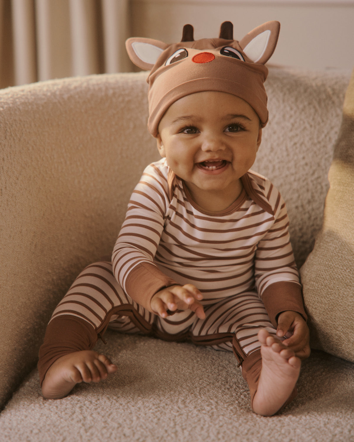 A baby wearing a brown and beige striped pajama suit and a matching reindeer hat with a red nose, sitting on a beige armchair.