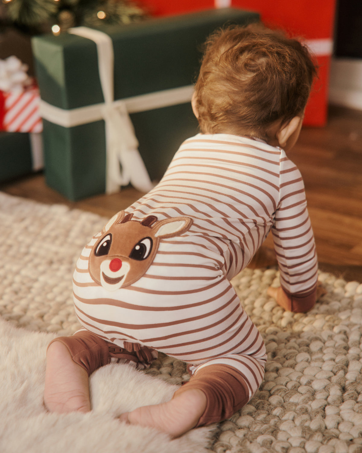A baby wearing a brown and cream striped long-sleeve footie with a Rudolph the Red-Nosed Reindeer face graphic on the back, crawling on a textured rug in a holiday setting.