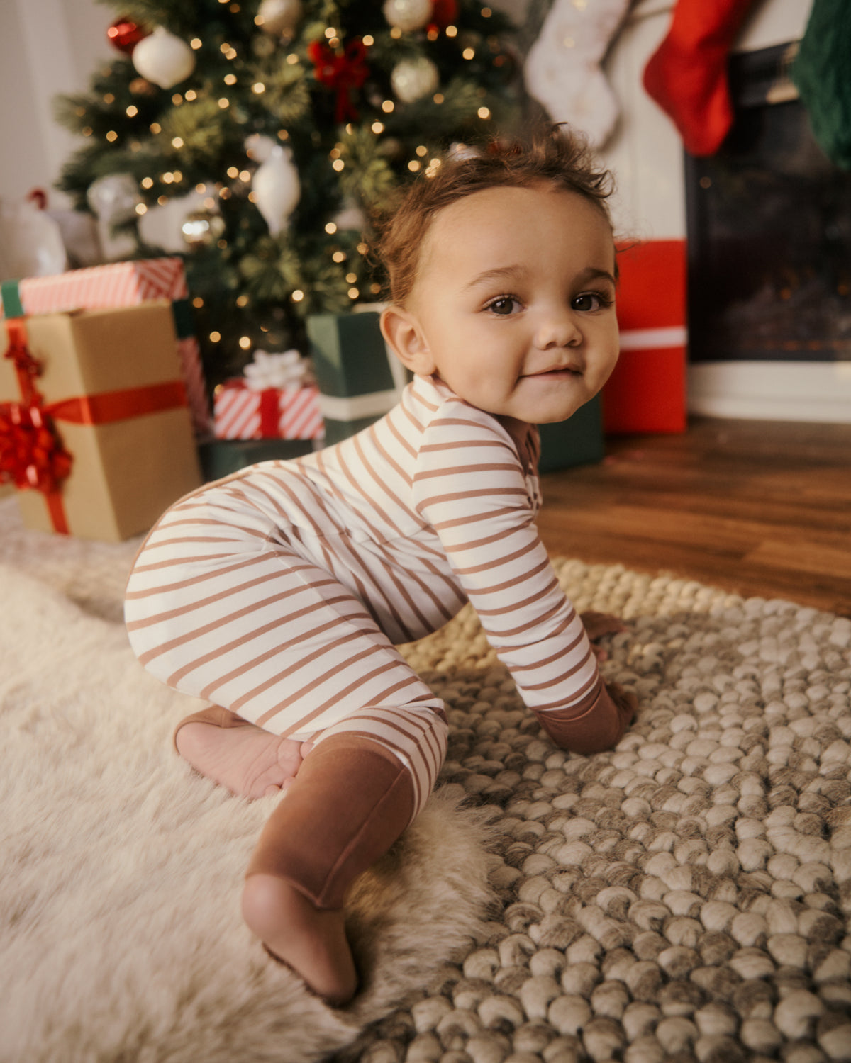 A baby wearing a cream and brown striped long-sleeve footie crawls on a textured rug in front of a decorated Christmas tree and fireplace.