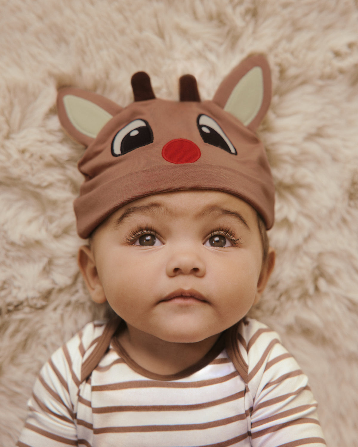 A baby wearing a brown reindeer-themed baby hat with antlers, large eyes, and a red nose, lying on a fluffy cream-colored blanket.