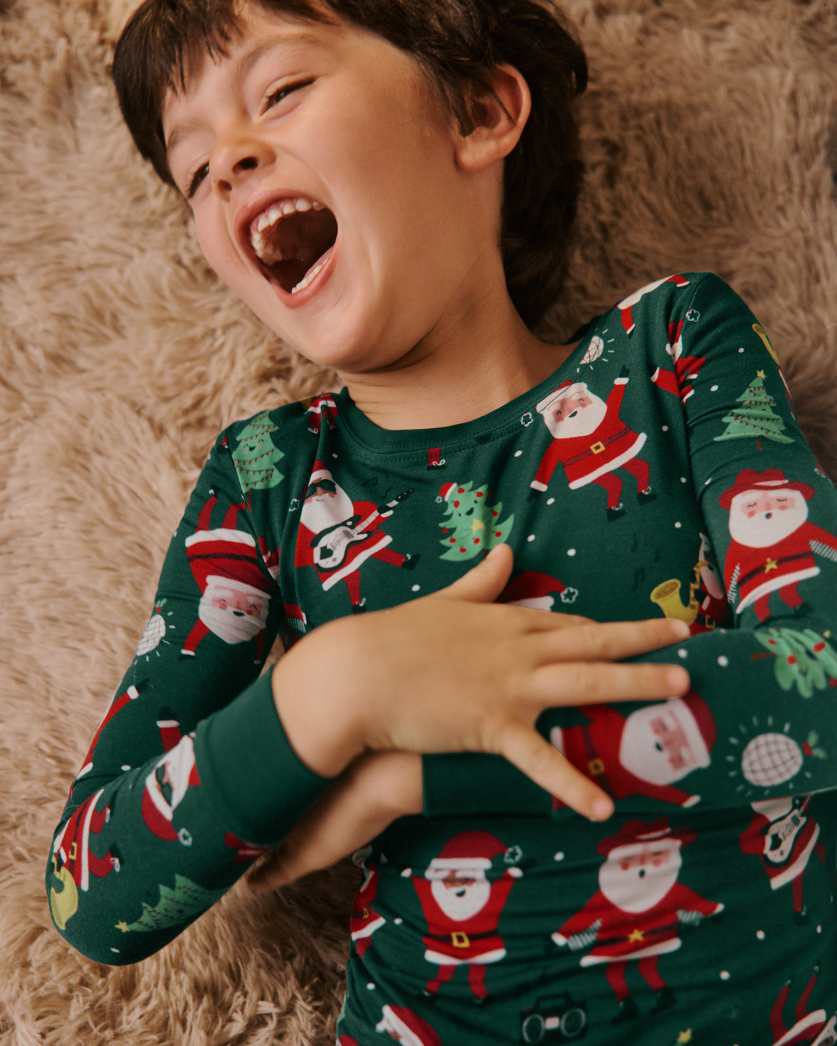 A young boy laughing on a brown shag rug, wearing a dark green long-sleeve pajama shirt with a festive Santa Claus print.