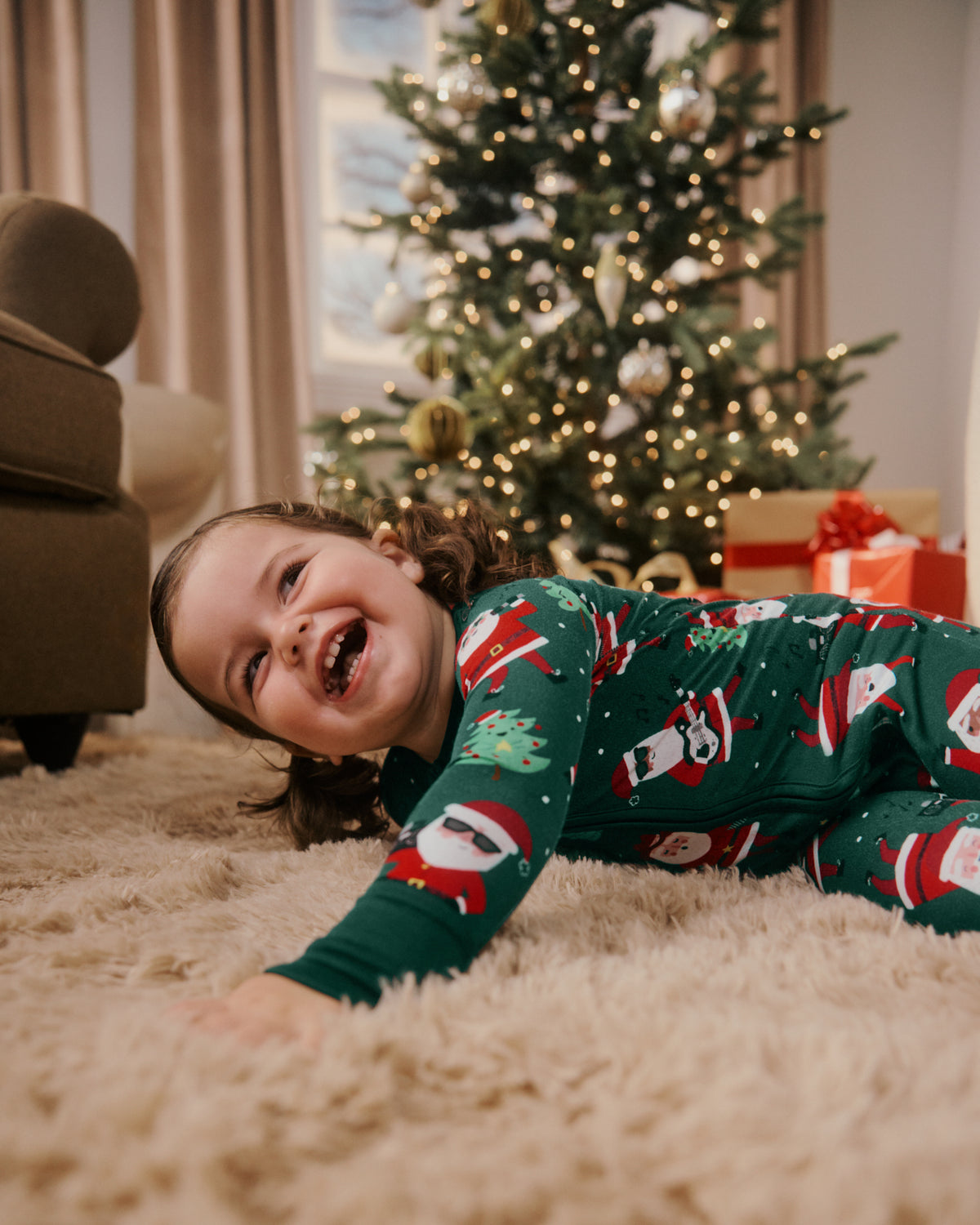 A young child wearing a green Christmas-themed pajama set with Santa Claus prints lies on a fluffy cream rug in front of a decorated Christmas tree and wrapped gifts.