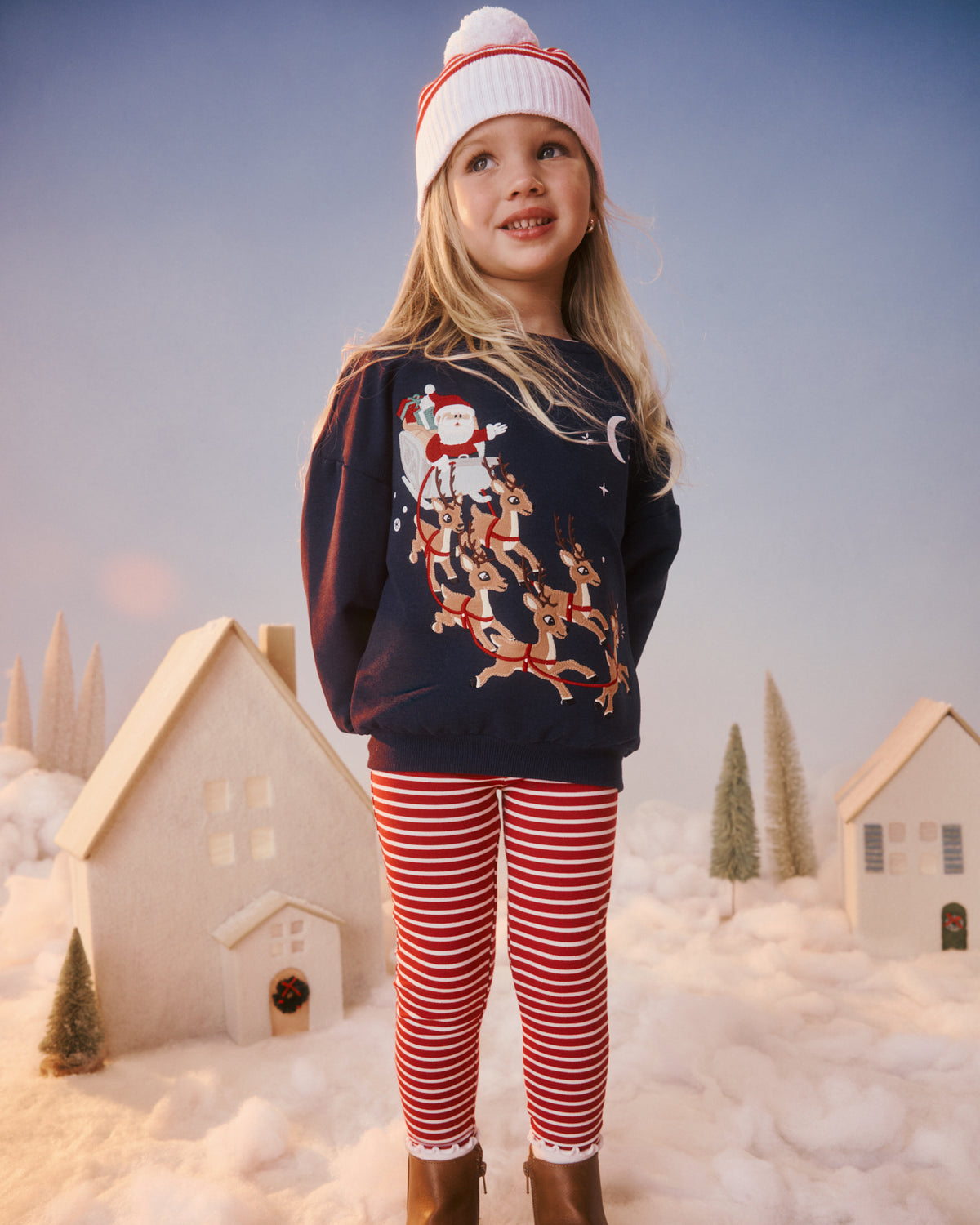 A young girl stands in a snowy village scene wearing a navy Christmas sweater with a festive graphic, red and white striped leggings, a white knit beanie with red stripes, and brown boots.
