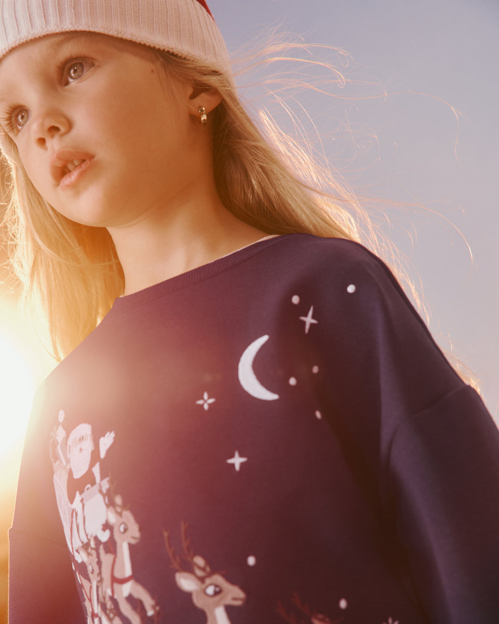 A young girl wearing a festive dark blue Christmas sweatshirt with white reindeer and moon graphics, paired with a white knit hat, captured in warm golden-hour sunlight.