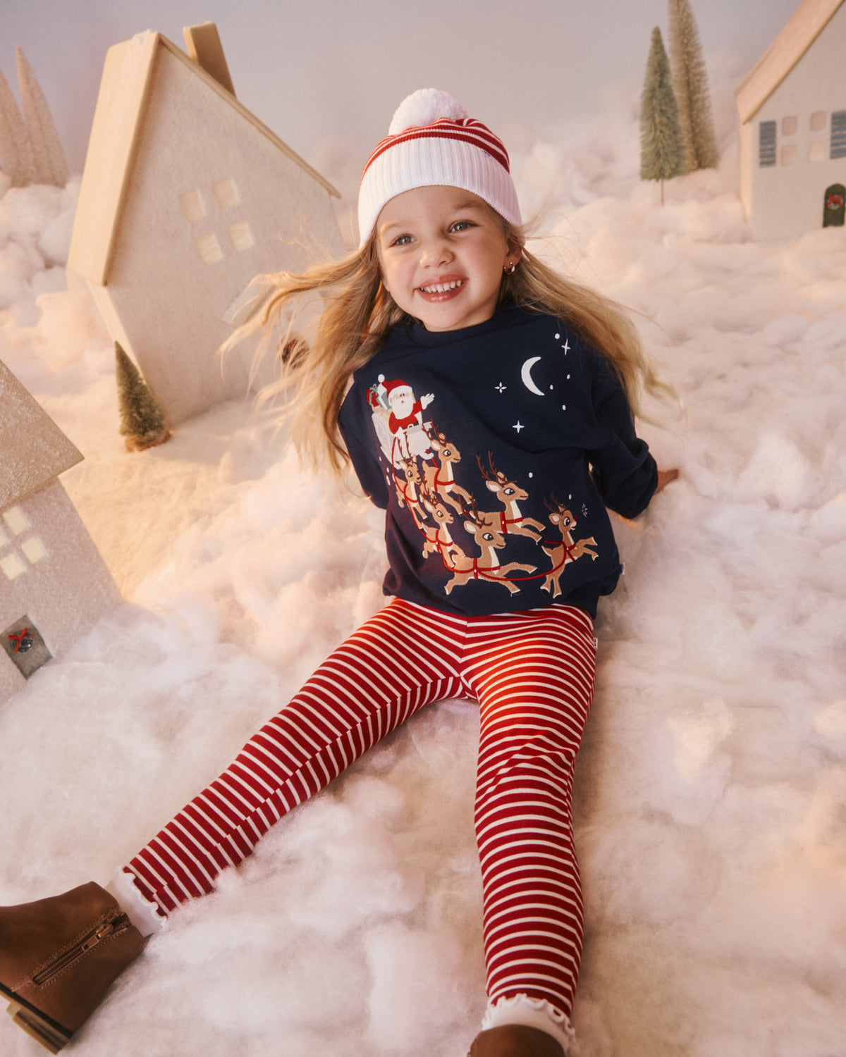 A young girl wearing a navy blue Christmas sweater with a Santa and reindeer graphic, red and white striped leggings, a white beanie with red stripes, and brown boots, sitting in a snowy, whimsical miniature village setting.