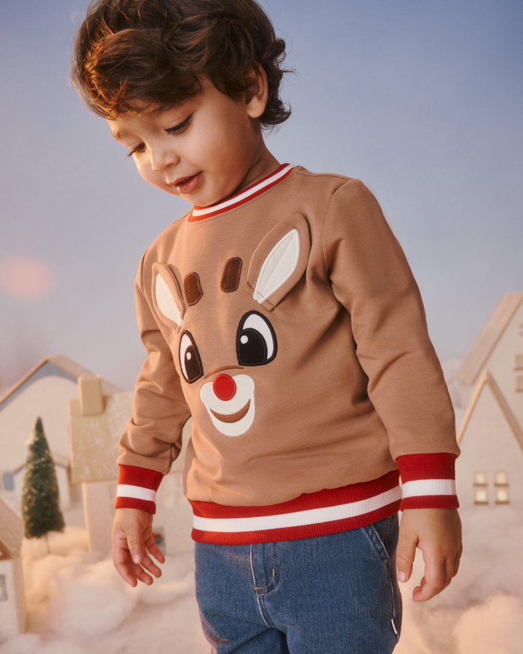A young boy wearing a brown long-sleeve sweater with a Rudolph the Red-Nosed Reindeer face graphic, styled with blue jeans in a whimsical, snowy village setting.