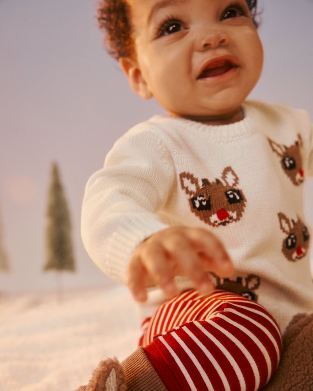A close-up lifestyle shot of a baby wearing a white knit sweater with brown reindeer faces and red-and-white striped pants, sitting in a cozy, softly lit setting.
