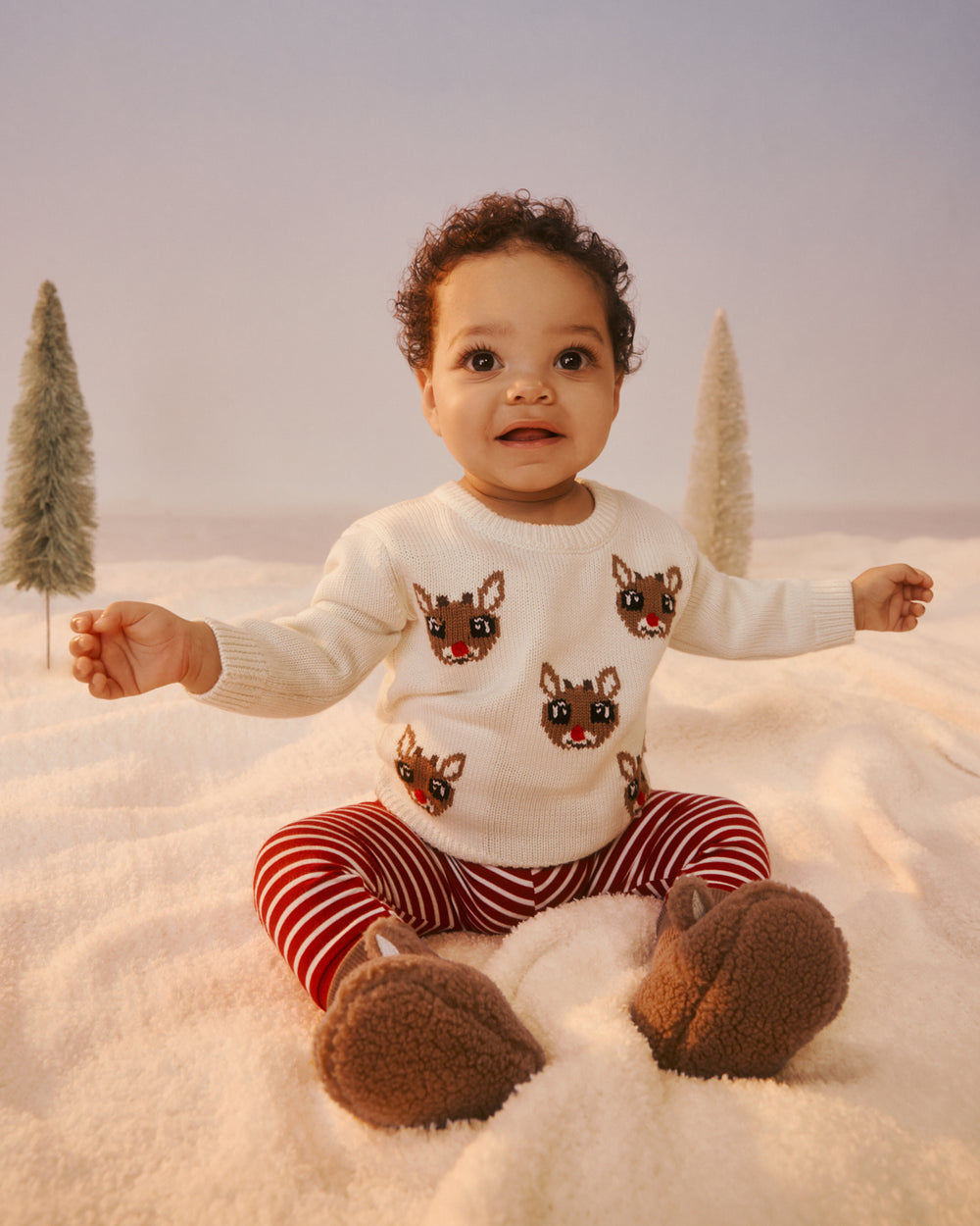 A baby sitting in a snowy studio setup wearing a white knit sweater with reindeer face prints, red and white striped pants, and brown fuzzy booties, with small decorative trees on either side.