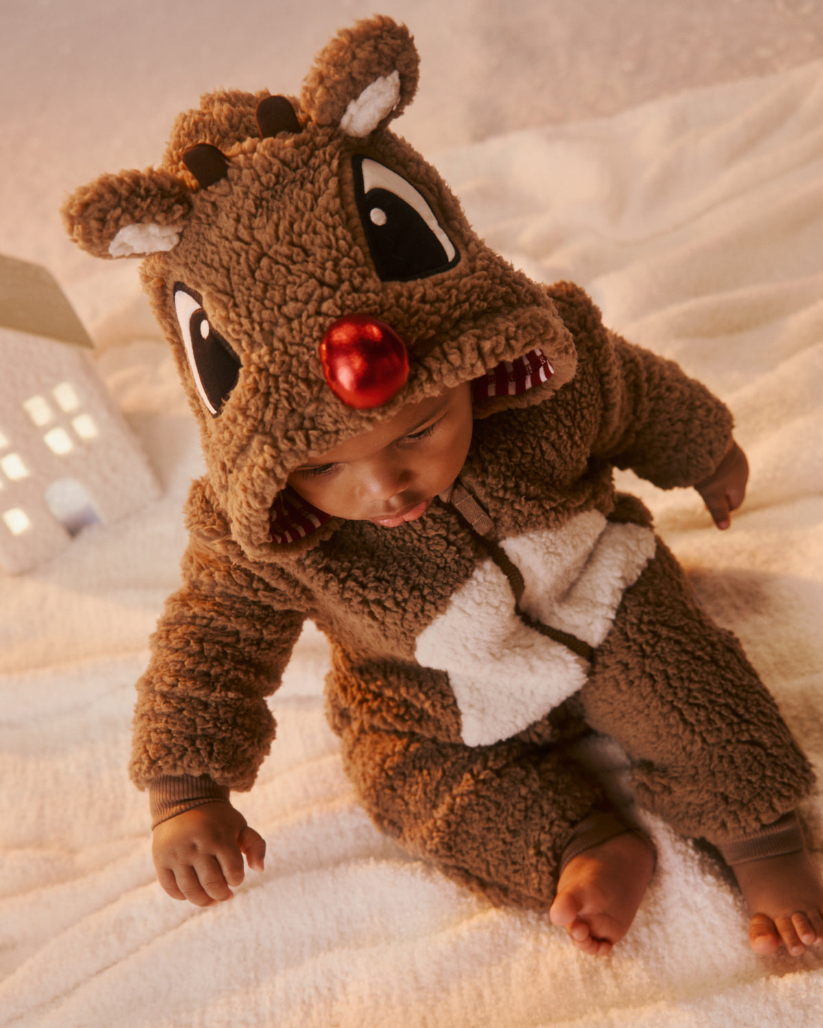 A baby wearing a brown plush reindeer romper with a red nose and white bow tie, sitting on a soft cream blanket.