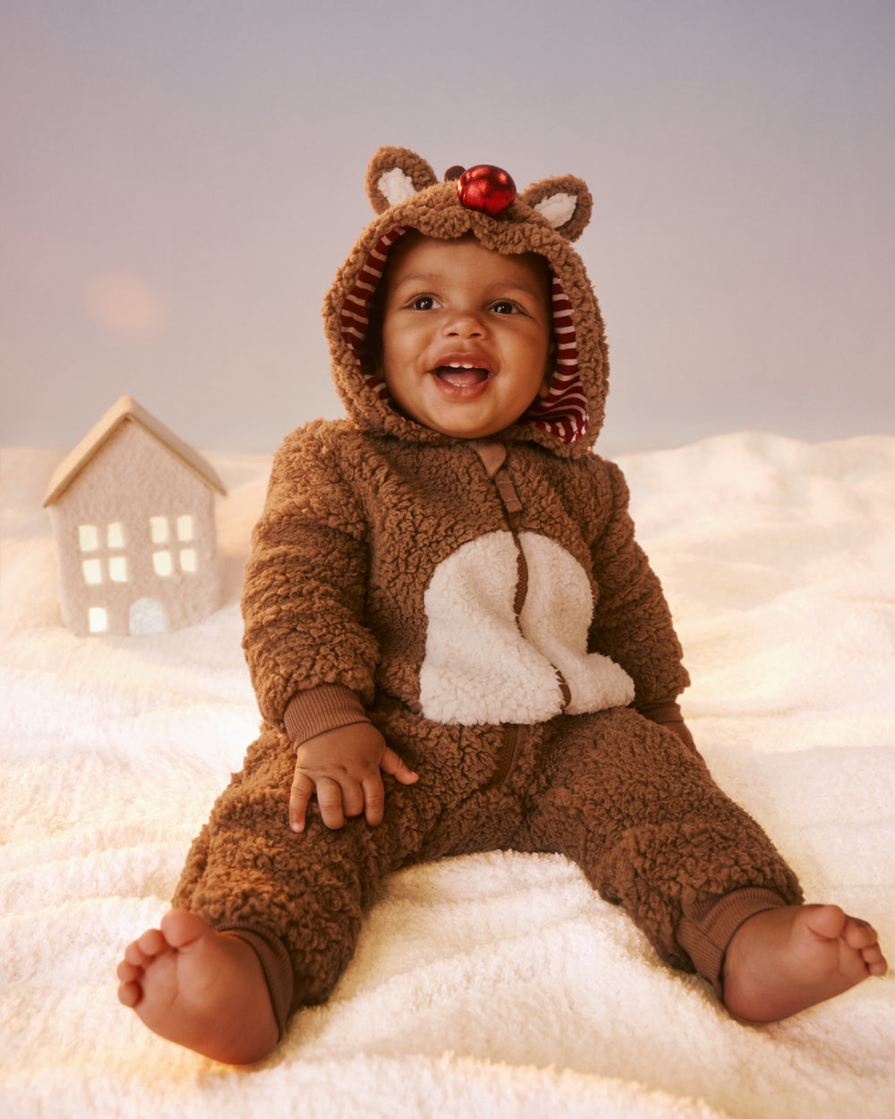 A baby wearing a brown sherpa romper with a white belly patch and a red ornament on the hood, sitting on a white blanket with a small house prop in the background.