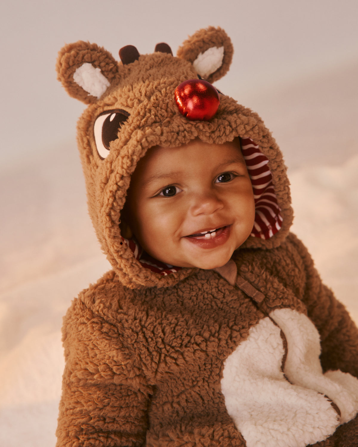 A baby wearing a brown plush reindeer costume with a hood featuring antlers, a red nose ornament, and a white belly patch, photographed against a soft beige background.