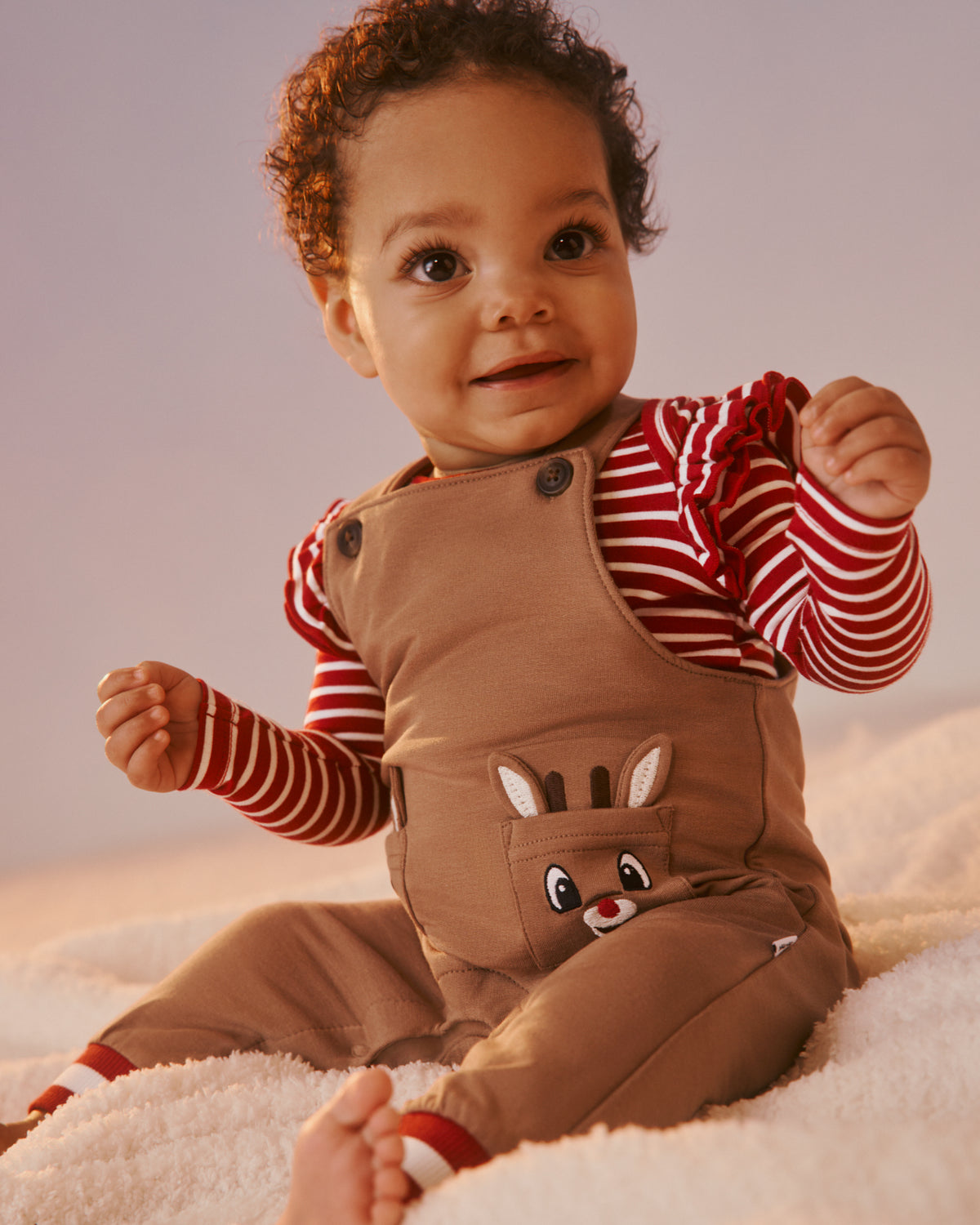 A baby sitting on a white textured blanket wearing a brown corduroy overall with a reindeer face patch on the pocket, layered over a red and white striped long-sleeve bodysuit.