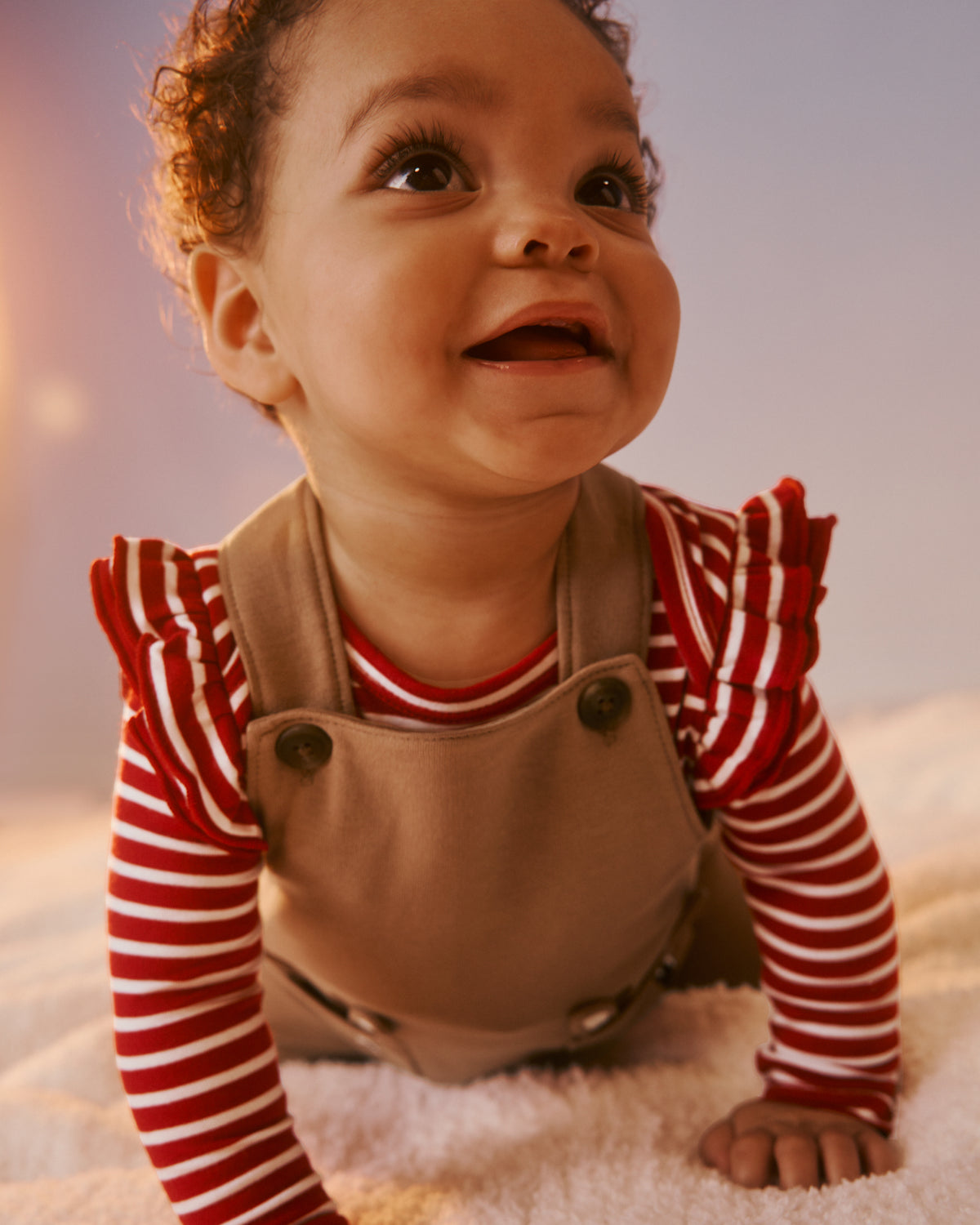A baby wearing a red and white striped long-sleeve shirt with ruffled shoulders and tan canvas overalls, crawling on a soft white blanket.
