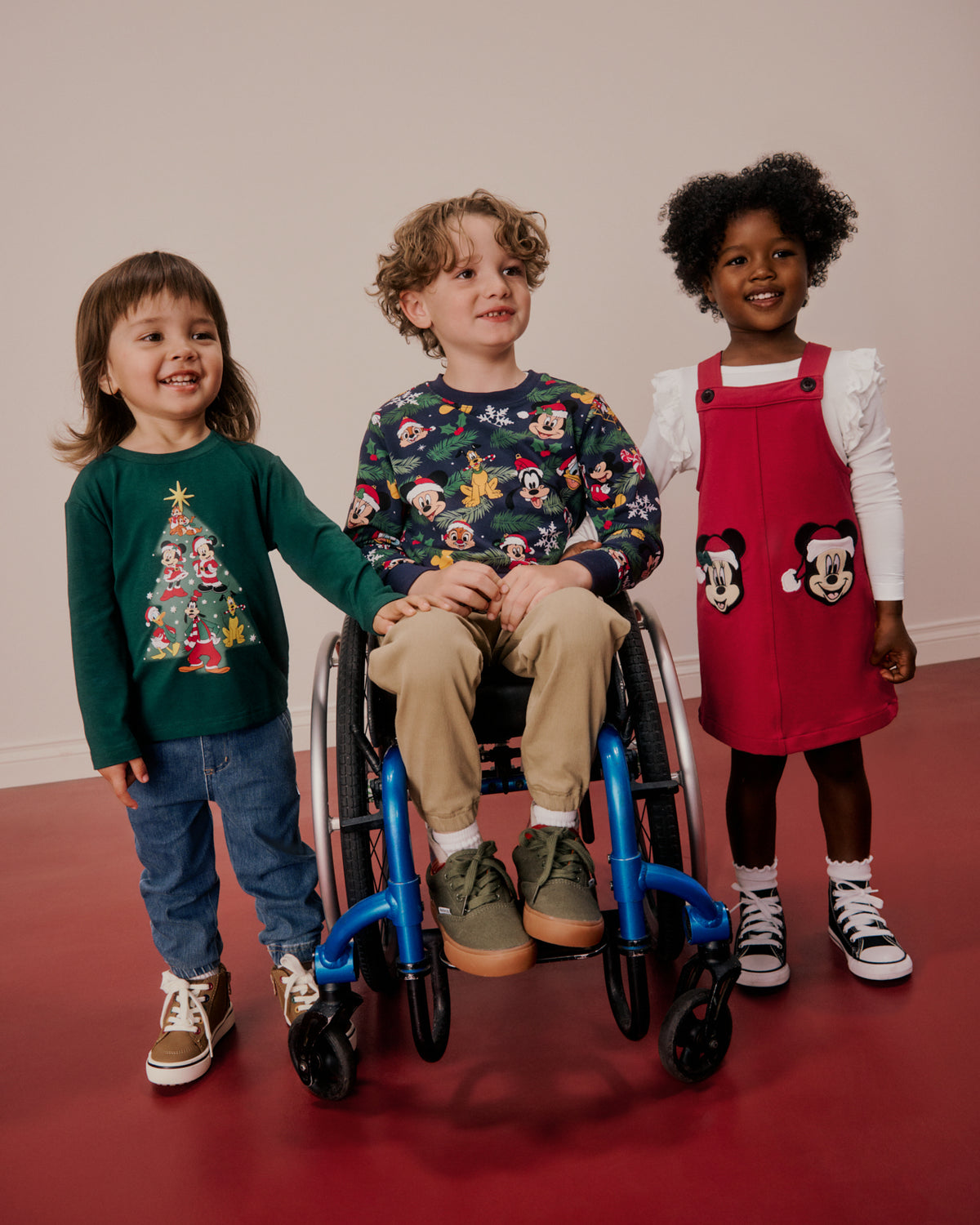 Three children standing indoors in a wheelchair and beside it, wearing holiday-themed apparel. The child on the left wears a dark green long-sleeve shirt with a Christmas tree graphic, blue jeans, and brown sneakers.