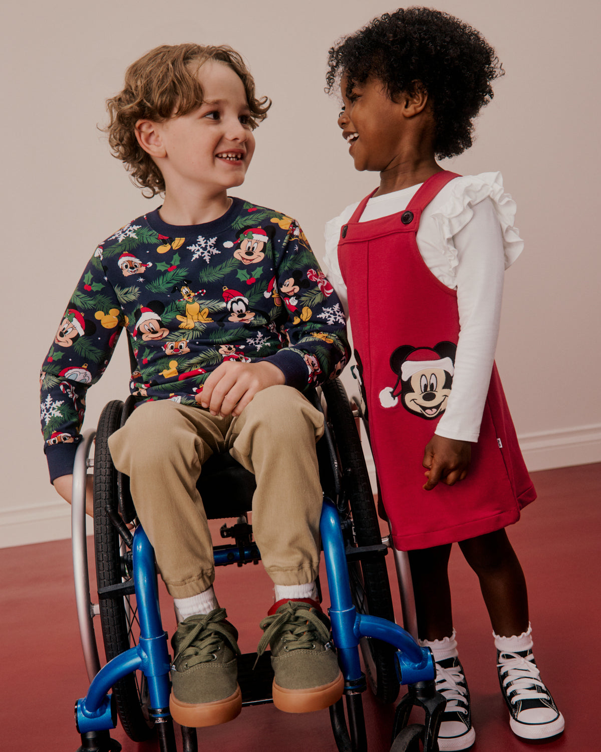 Two children wearing Disney Mickey-themed clothing, one sitting in a wheelchair and the other standing, photographed against a plain indoor wall with a red floor.