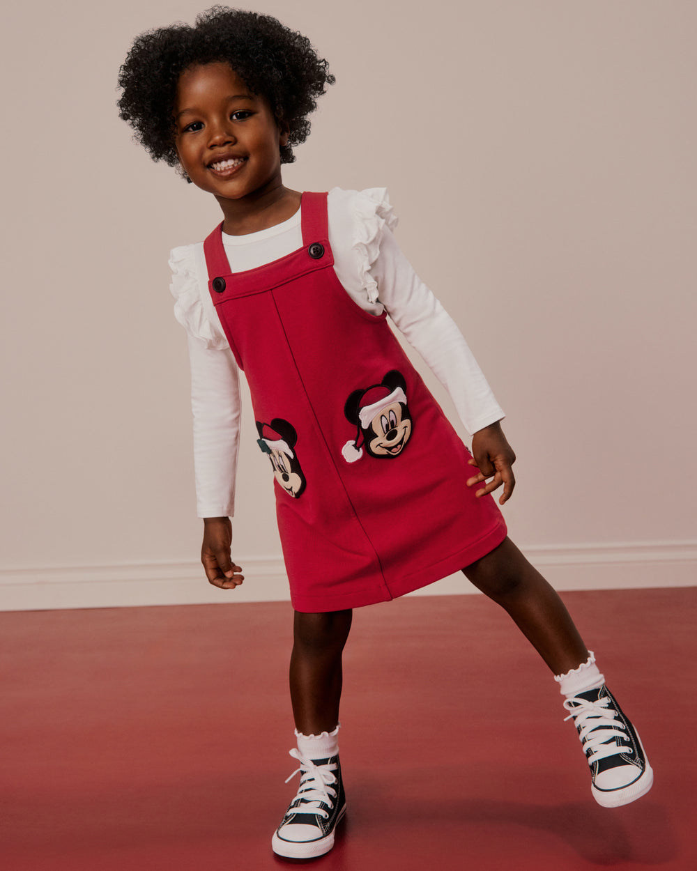 A young child wearing a red Minnie Mouse-themed pinafore dress over a white long-sleeve shirt, styled with black and white high-top sneakers, standing against a neutral beige wall and red floor.