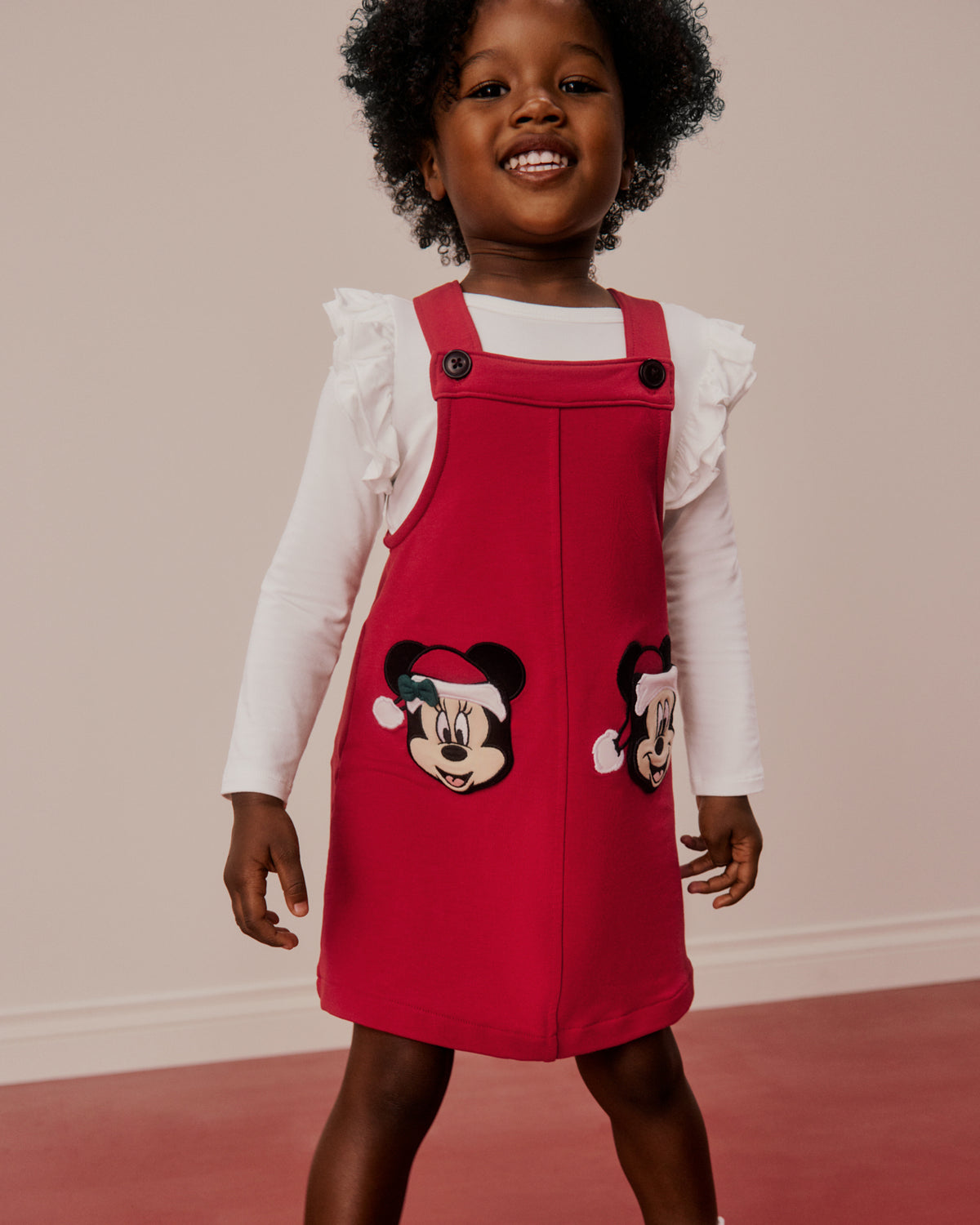 A young child wearing a red pinafore dress over a white ruffled long-sleeve shirt. The red dress features embroidered cartoon mouse faces on the front and side, set against a plain beige wall and pinkish-red floor.