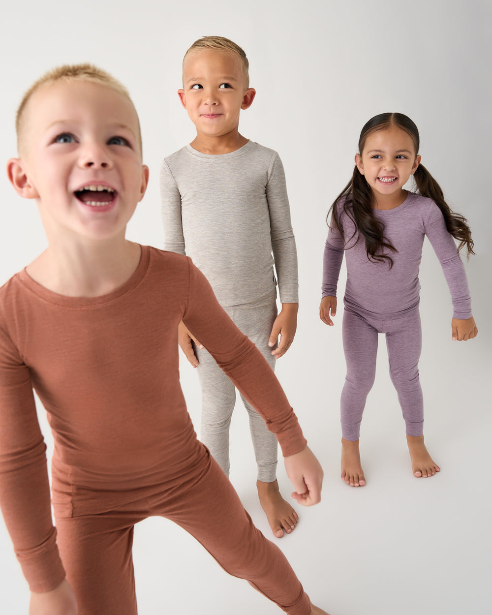 Three children modeling long-sleeve knit pajama sets in a studio setting. The child in the foreground wears a rust-brown set, the middle child wears a light gray set, and the child on the right wears a dusty lavender-purple set.
