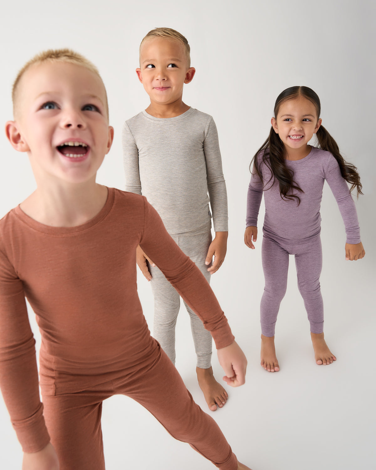 Three children modeling long-sleeve knit pajama sets in a studio setting. The child in the foreground wears a rust-brown set, the middle child wears a light gray set, and the child on the right wears a dusty lavender-purple set.