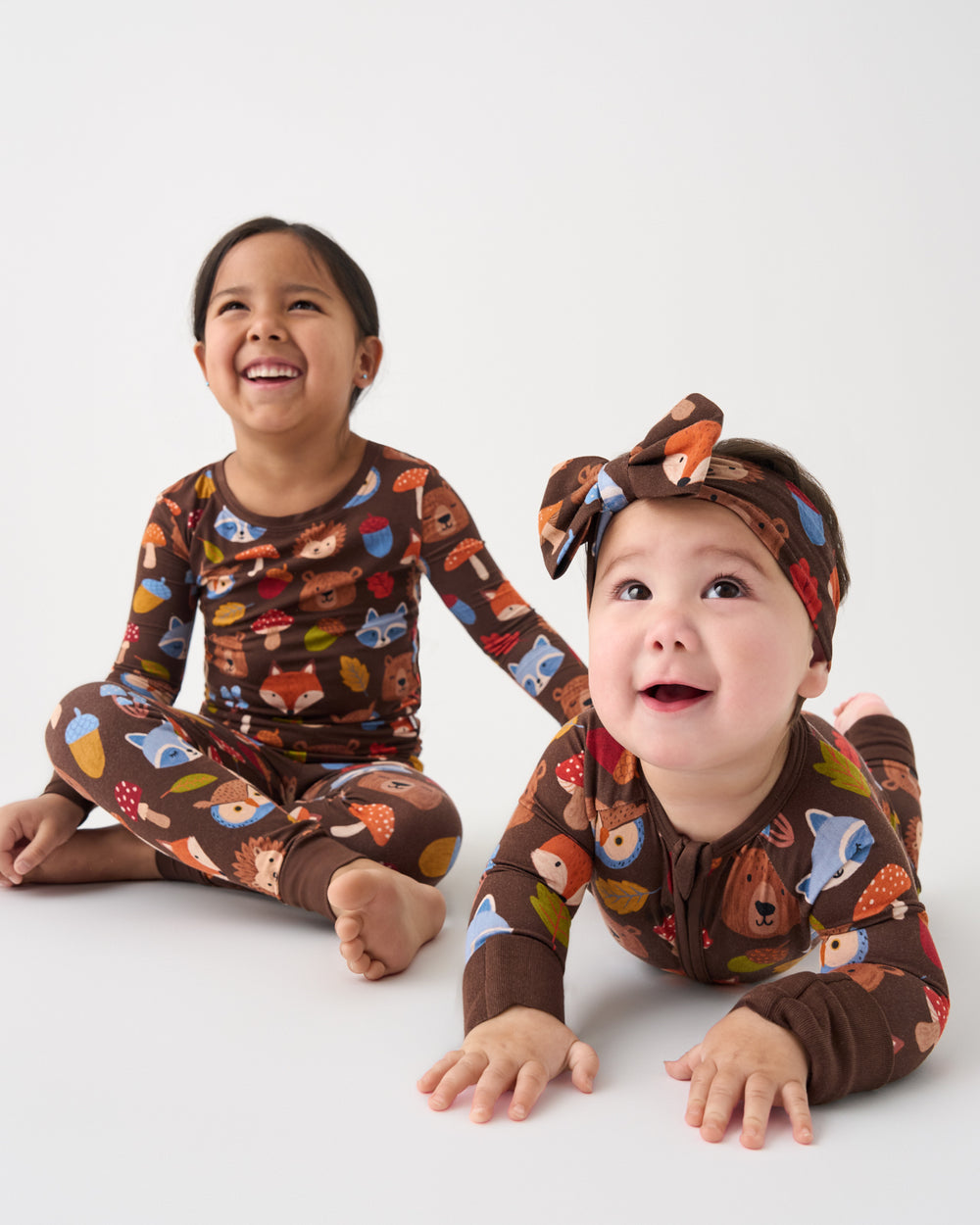 Two children wearing matching brown long-sleeve pajamas with a colorful woodland animal print, including a girl sitting cross-legged and a baby crawling, both with matching bow headbands, against a plain white studio background.