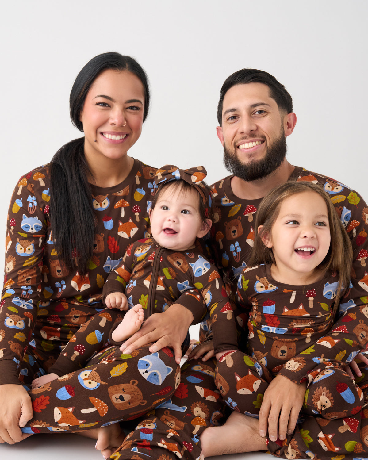 A family of four wearing matching brown long-sleeve pajamas with a colorful woodland animal print, including foxes, bears, deer, and mushrooms, posed together against a light gray studio background.