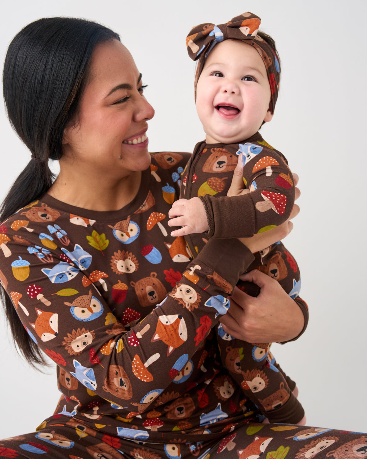 A smiling woman and a baby wearing matching brown pajamas with a colorful woodland print of hedgehogs, mushrooms, acorns, and leaves. The woman holds the baby, who is wearing a matching headband with a bow.