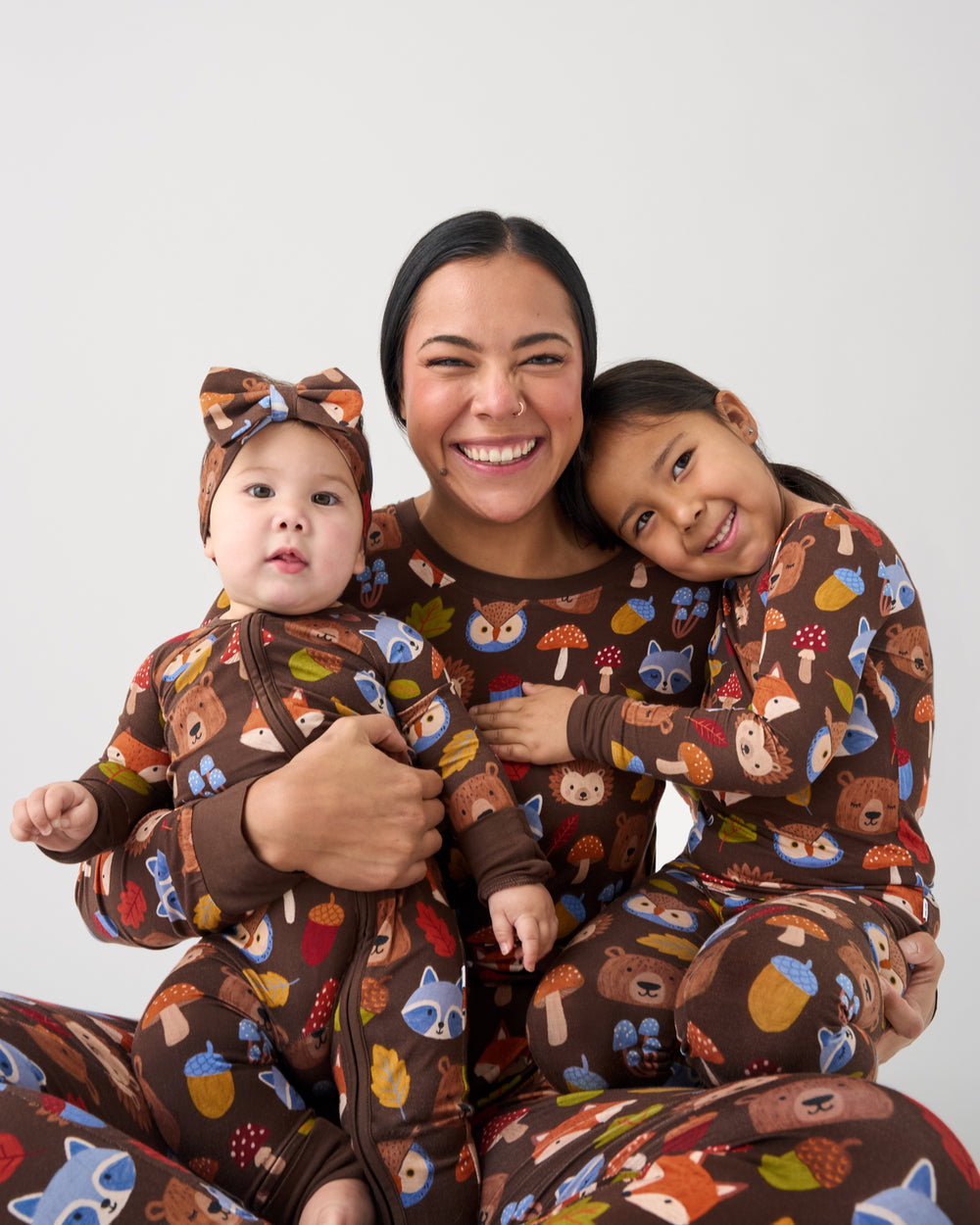 A smiling woman and two children wearing matching brown pajamas with a colorful woodland animal print, posed together against a light gray studio background.