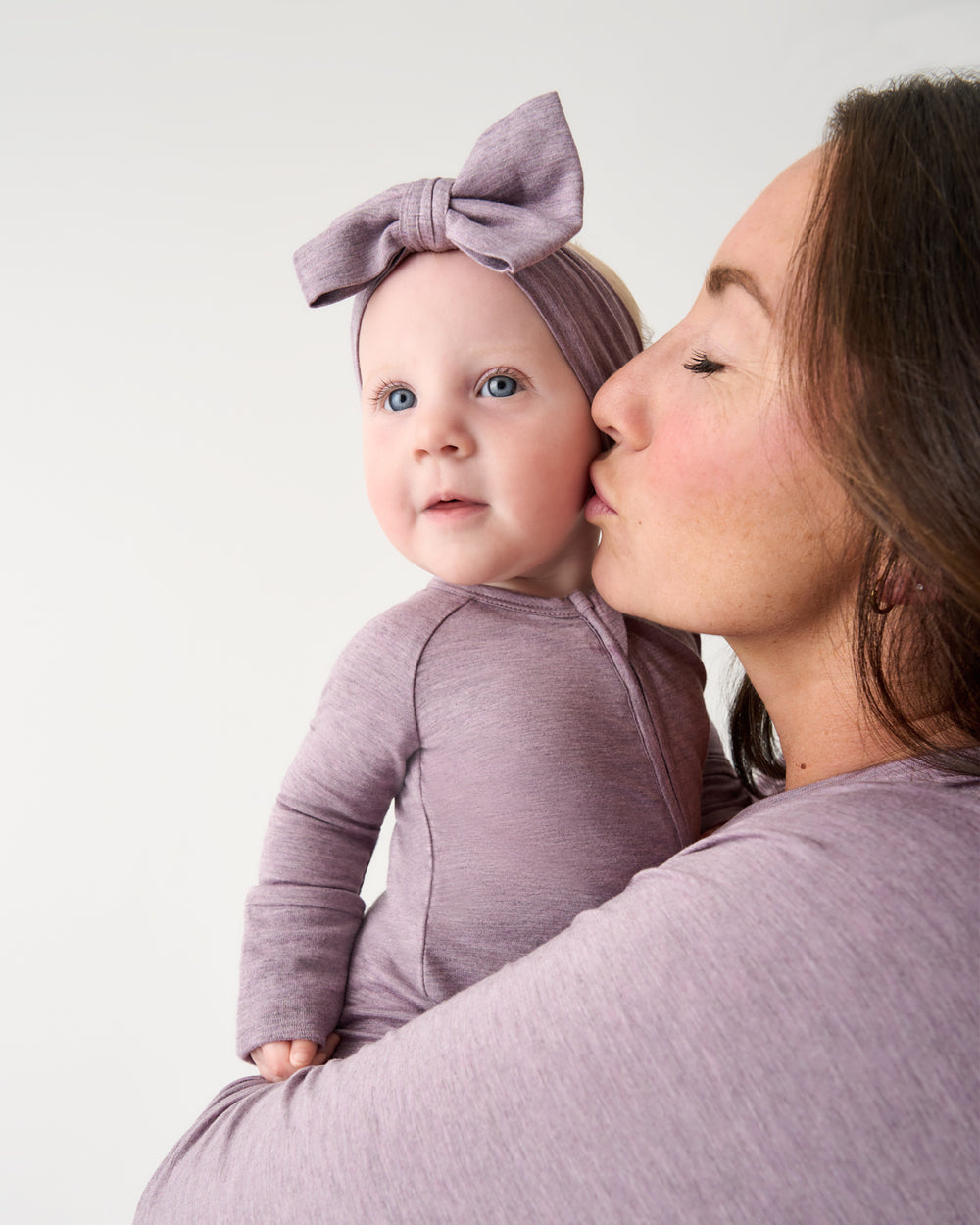 A woman holding a baby wearing matching mauve-purple long-sleeve bodysuit, headband with bow, and kissing the baby on the cheek against a plain light background.